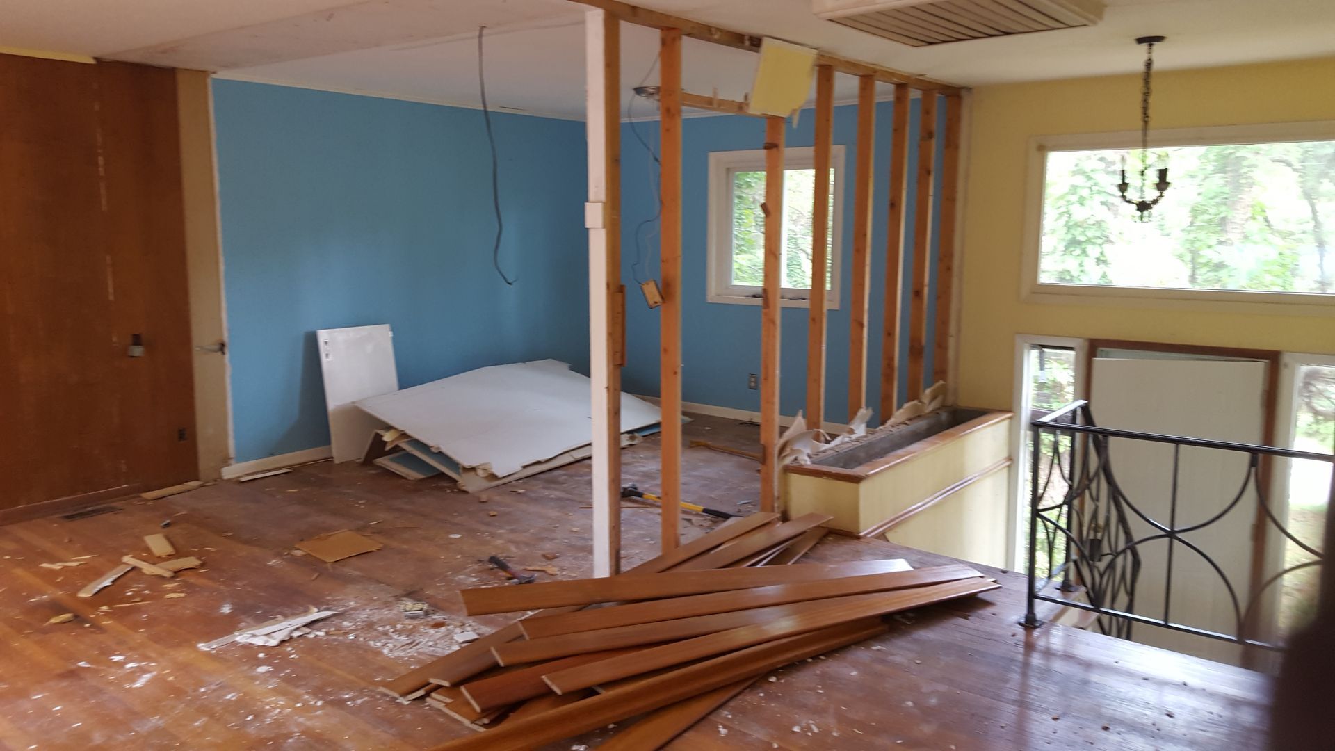A room under renovation with exposed wooden wall studs, blue painted walls, scattered debris, and an open stairwell.