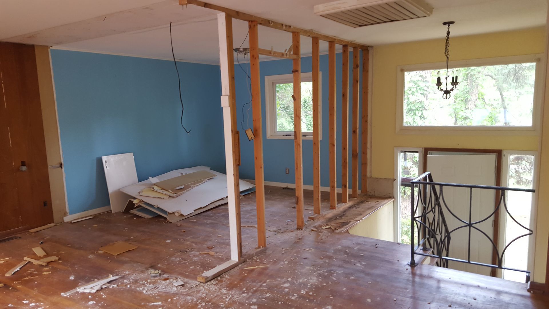 A room under renovation with exposed wooden wall studs, blue and yellow painted walls, debris on floor, and a staircase.