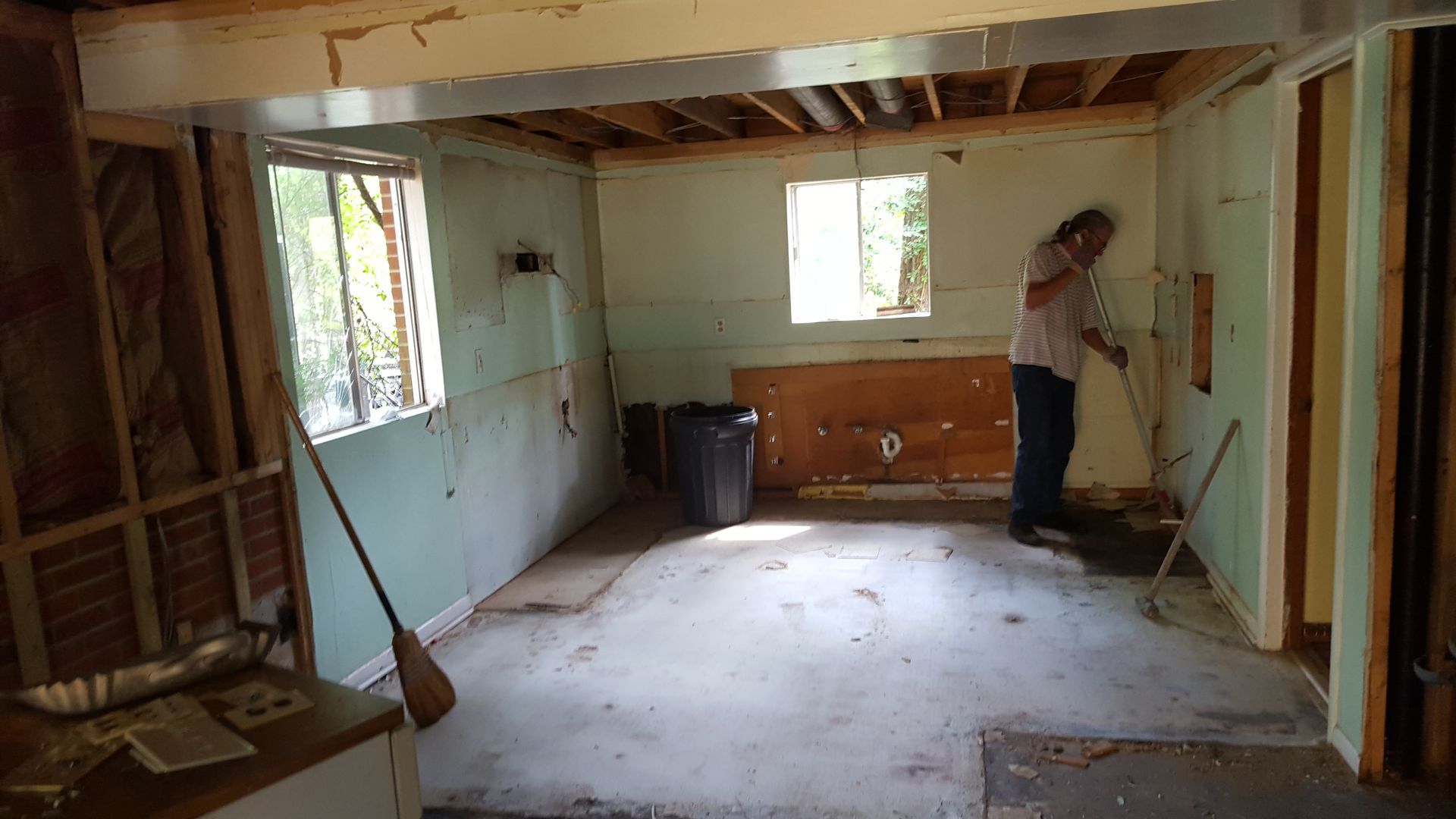 A person sweeps debris on a concrete floor in a room undergoing construction with exposed wall studs and framed windows.