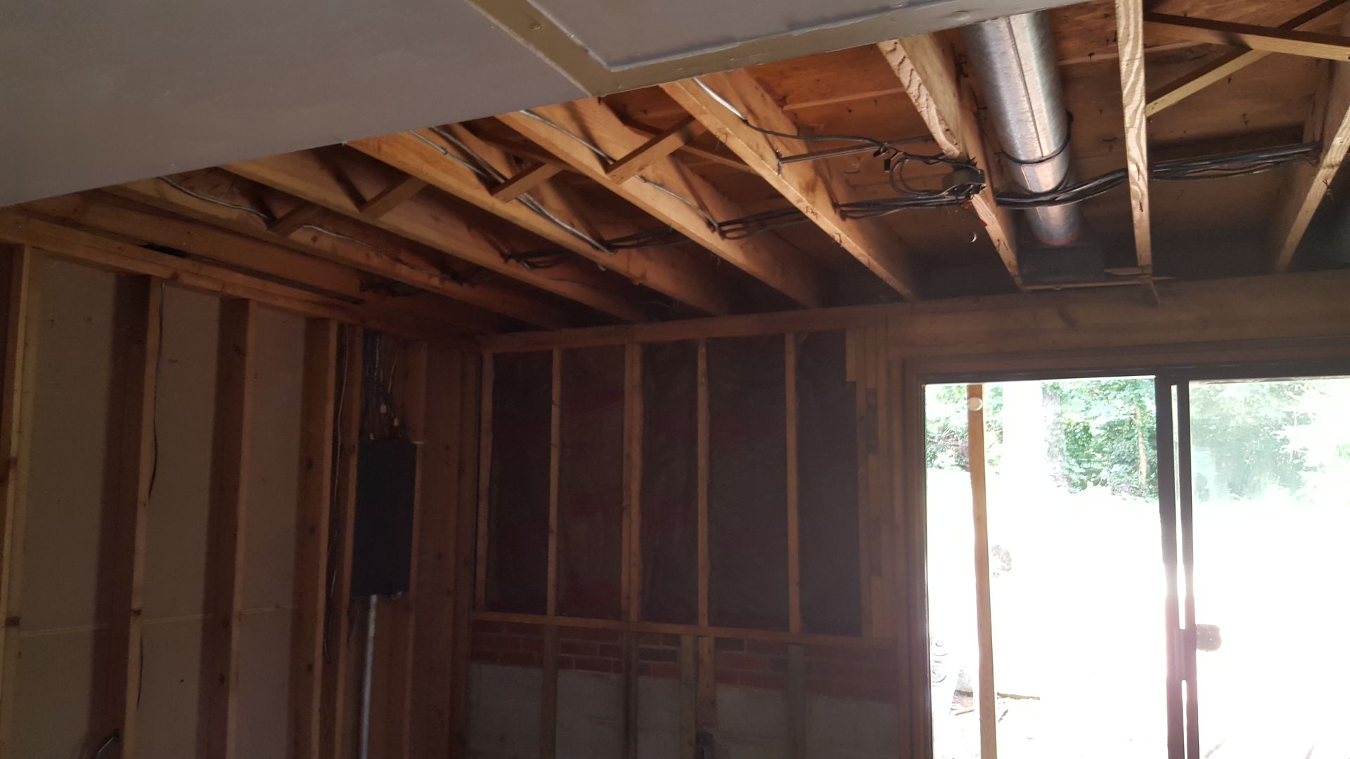 Interior view of an unfinished room showing exposed wooden ceiling joists, wall studs, a metal duct, and a glass door.