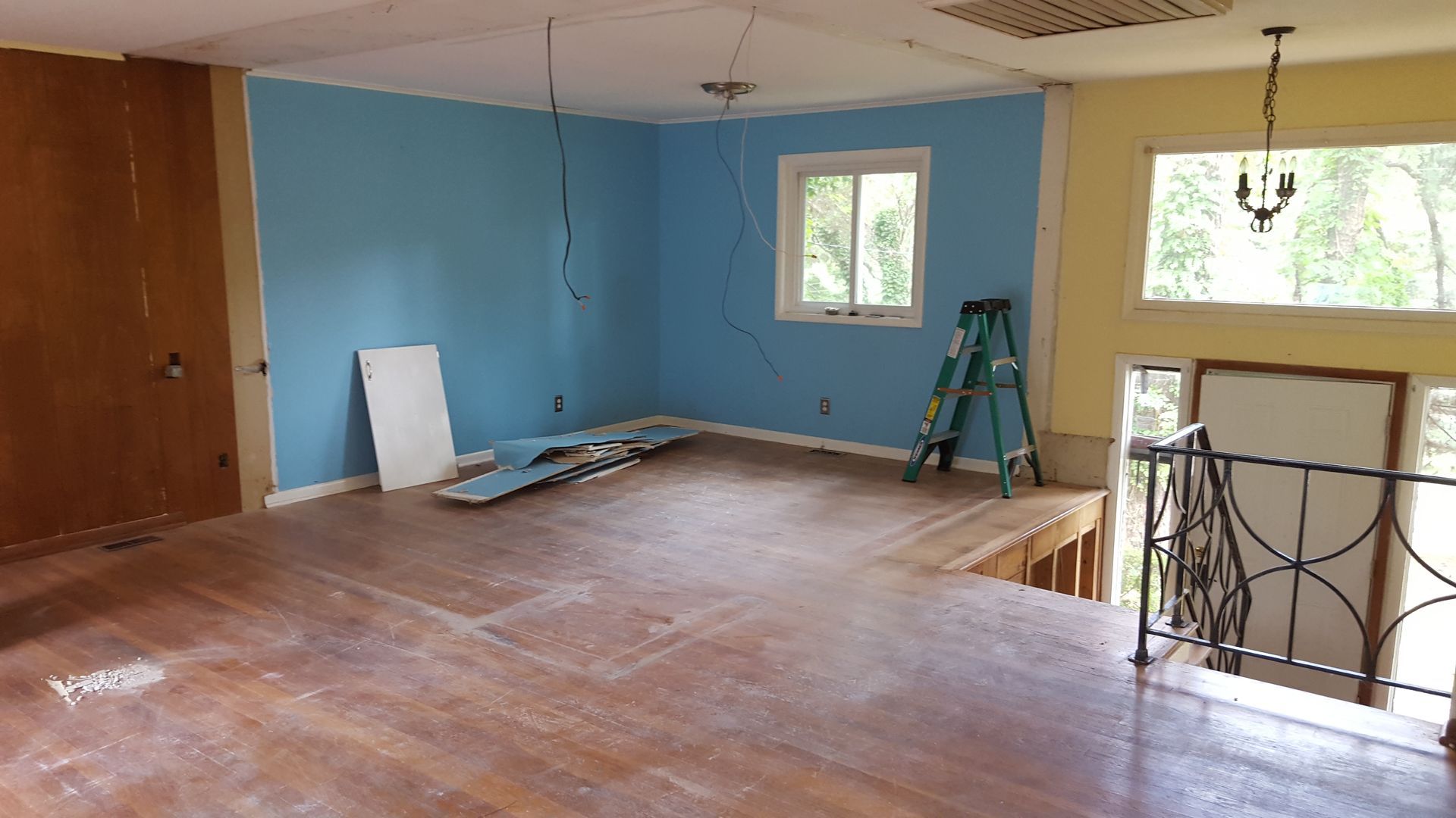 A room under renovation with blue and yellow walls, exposed wood floors, a stepladder, and an open staircase.