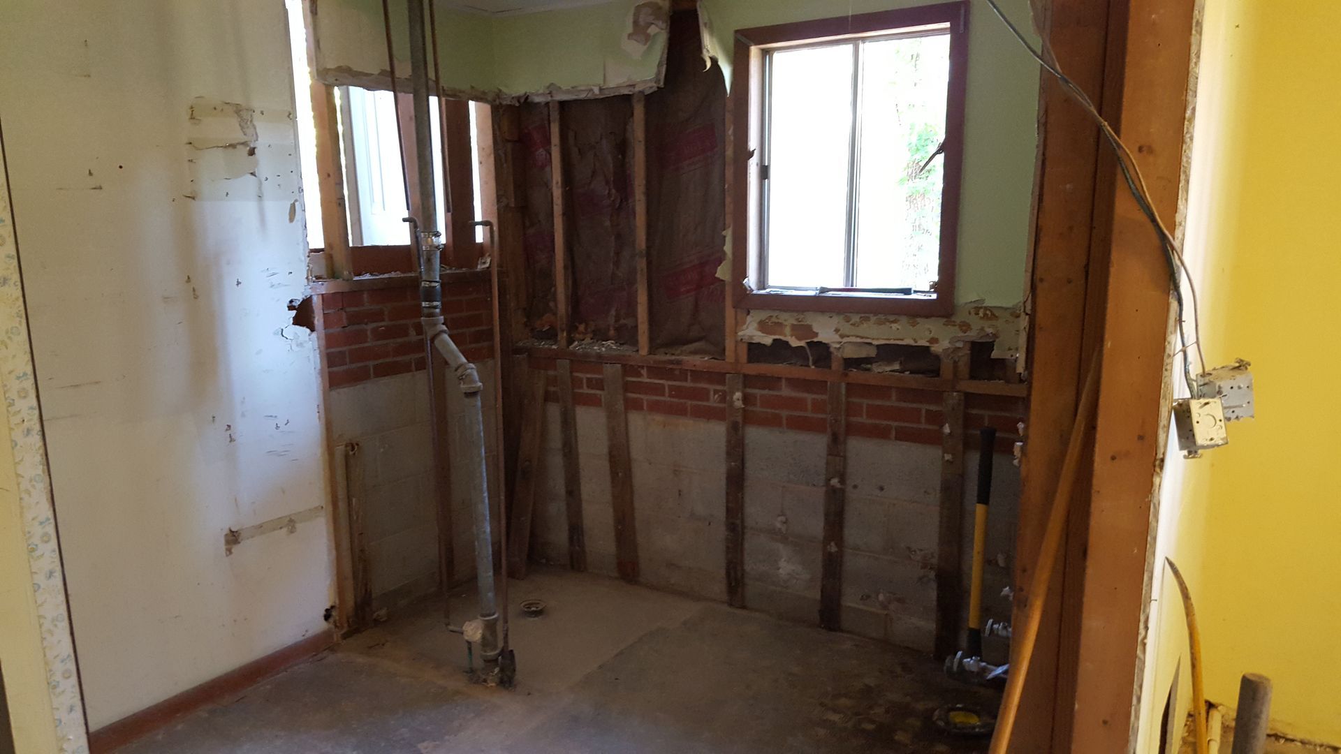 A room under renovation with exposed wooden studs, brick walls, and a window, with plumbing pipes visible on the floor.