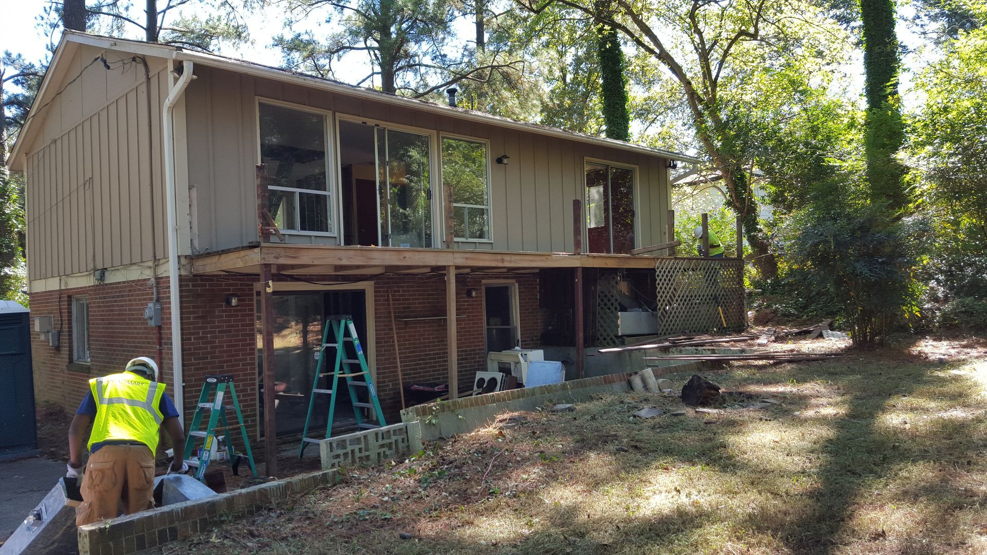 A worker in a high-visibility vest stands near a ladder while renovating the deck of a two-story house.