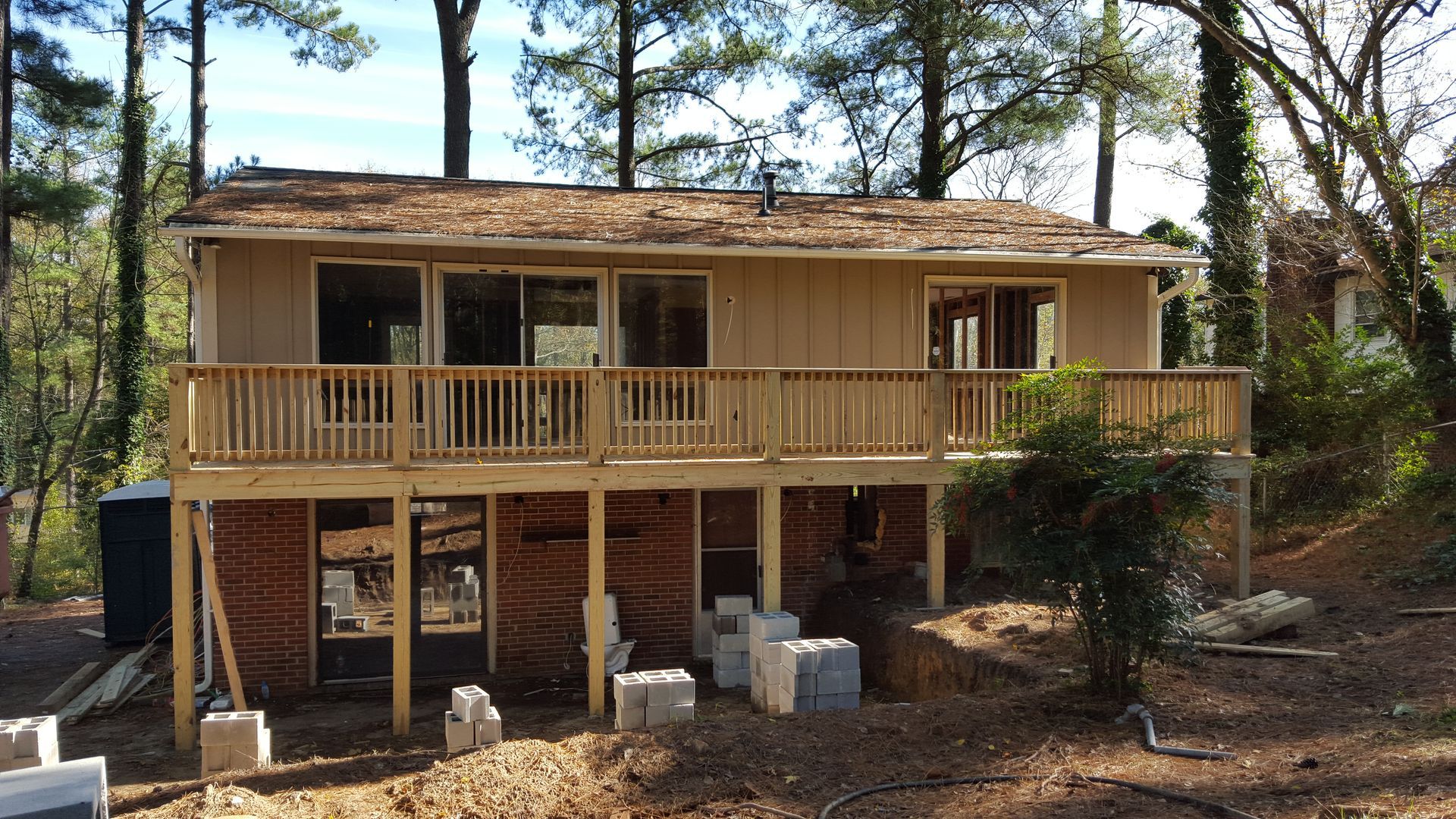 A tan house with a new wooden deck under construction, surrounded by trees and a dirt yard with concrete blocks.