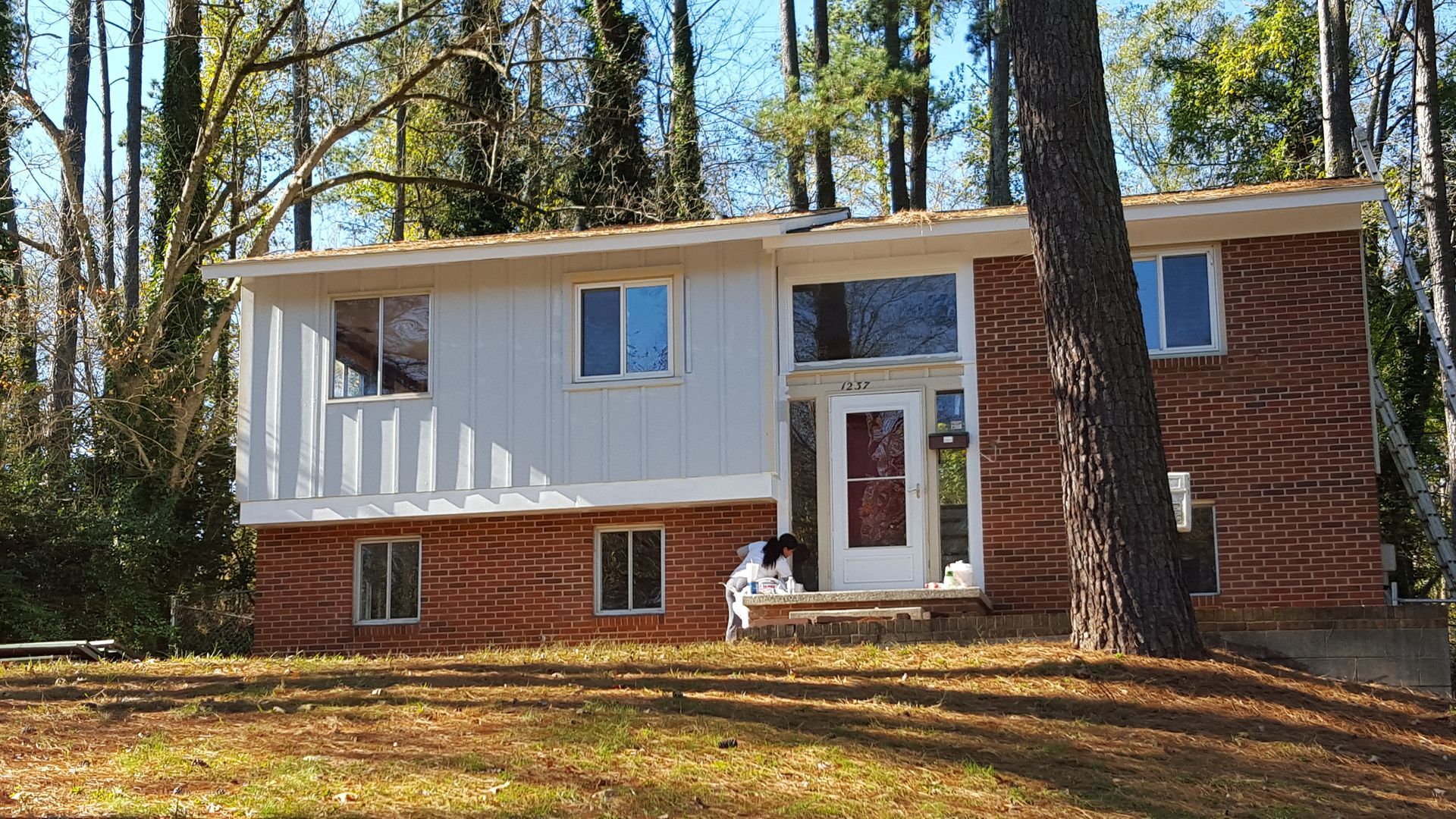 A two-story split-level home with a red brick lower level, grey vertical siding on the upper left, and a white front door.