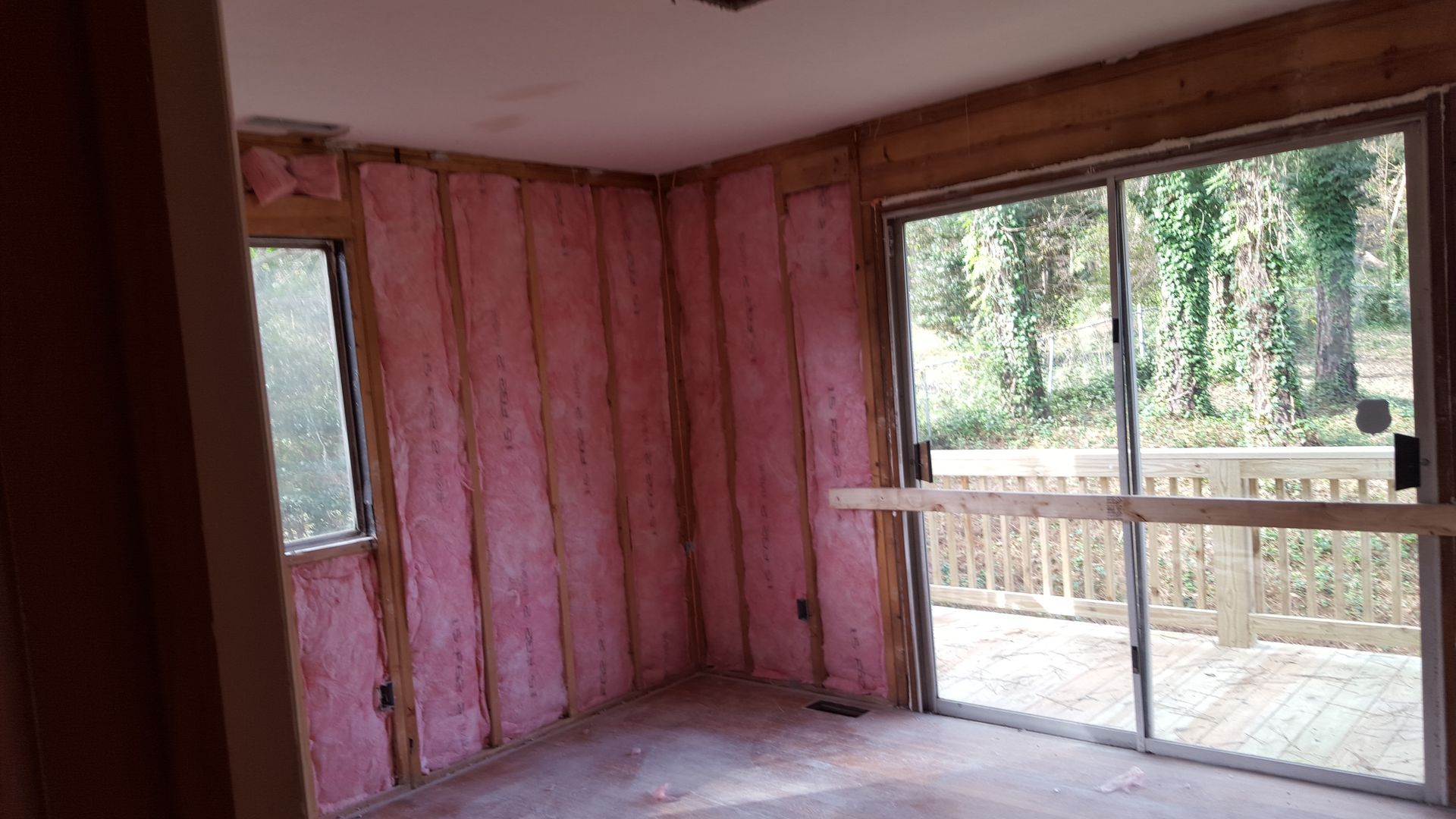A room under renovation with pink fiberglass insulation installed between wall studs, a side window, and a sliding door.