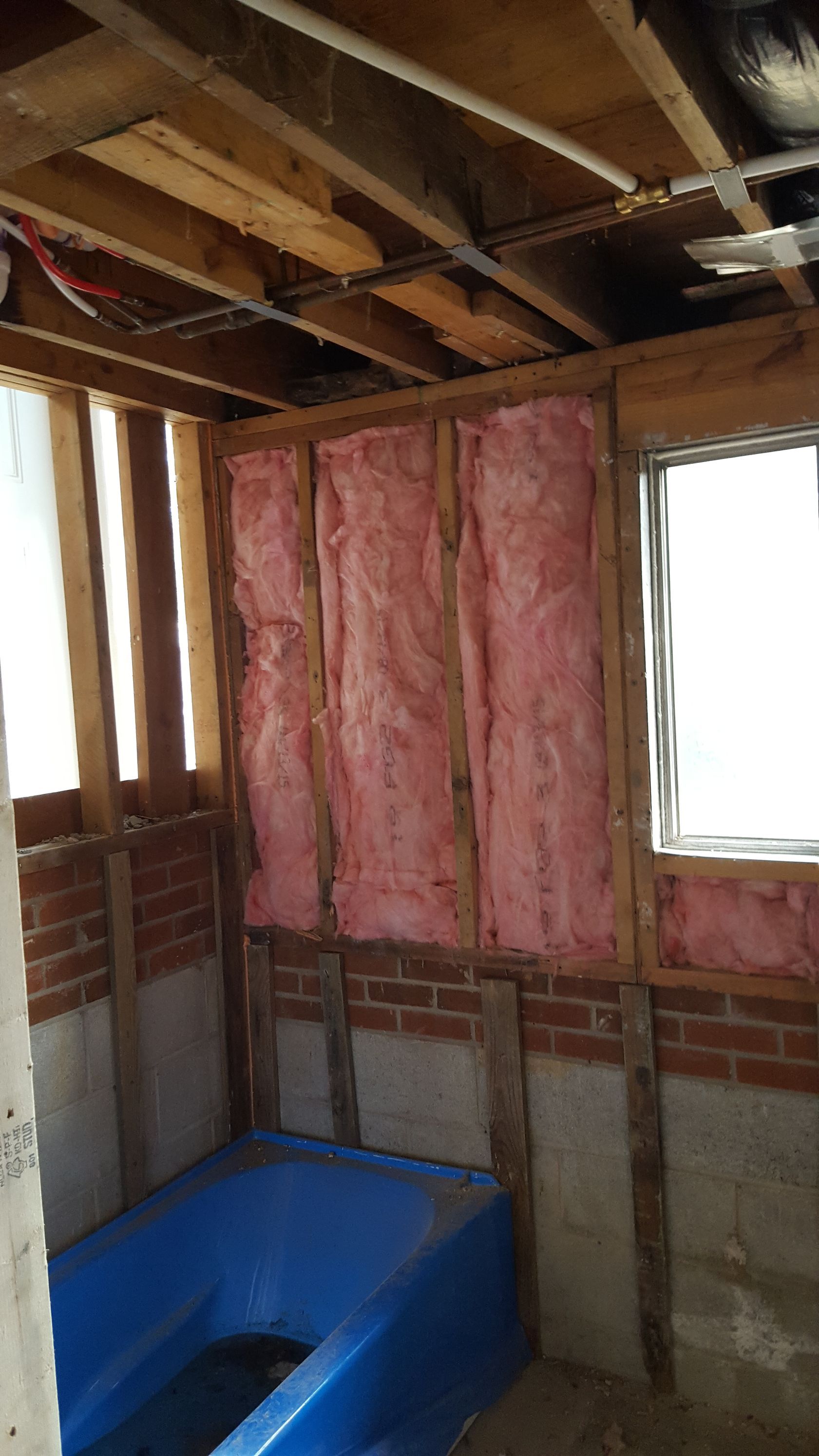 An unfinished bathroom with a blue bathtub, exposed wooden framing, brick foundation, and pink fiberglass insulation.