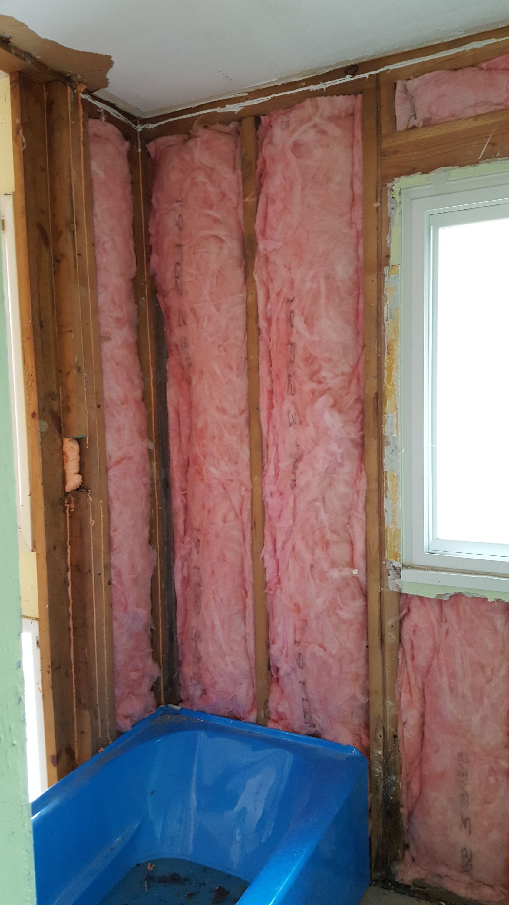 A blue bathtub sits in an unfinished bathroom alcove with exposed wall studs and pink fiberglass insulation.
