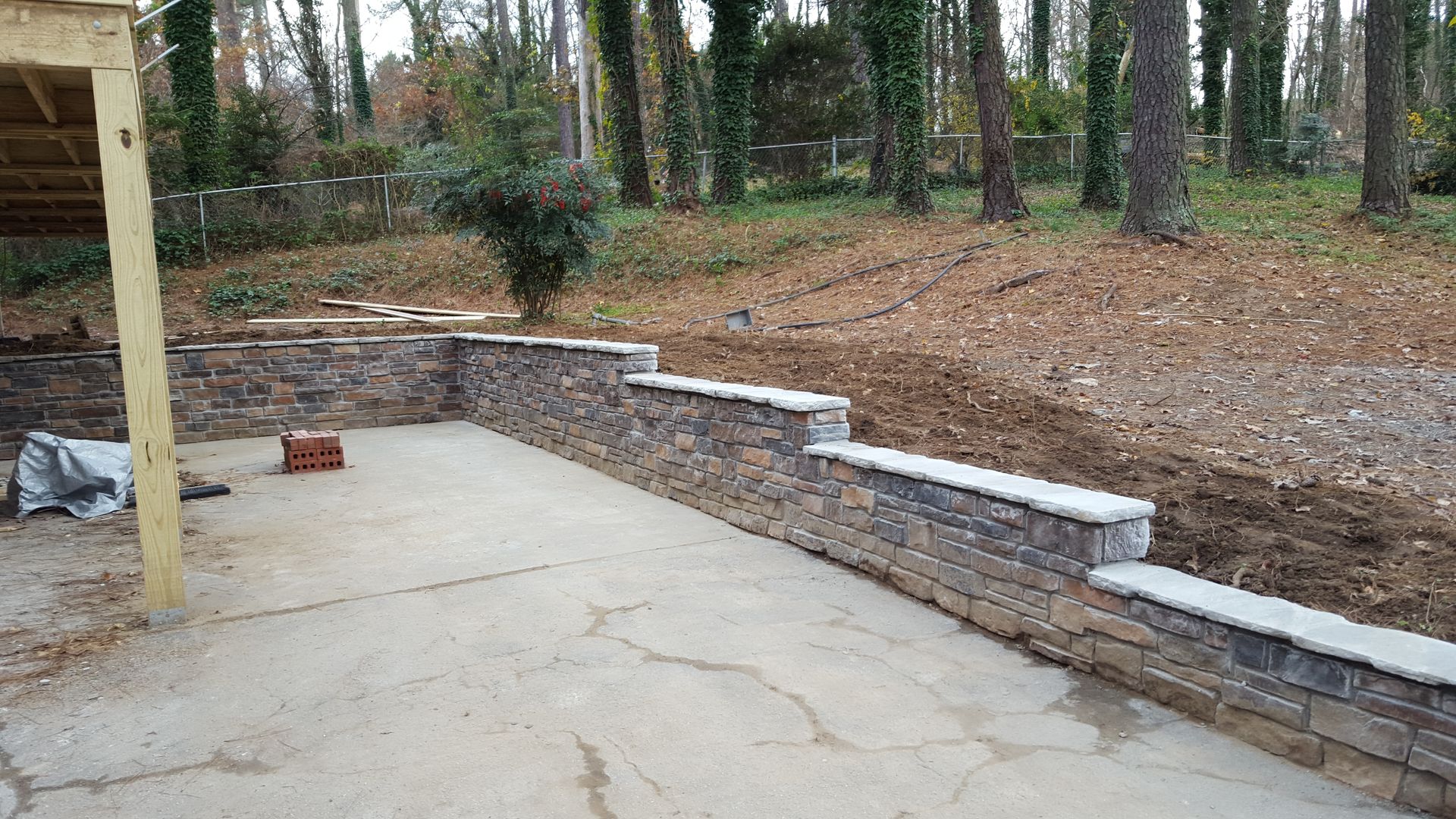 A tiered, stone retaining wall borders a concrete patio in a yard with trees and exposed soil in the background.