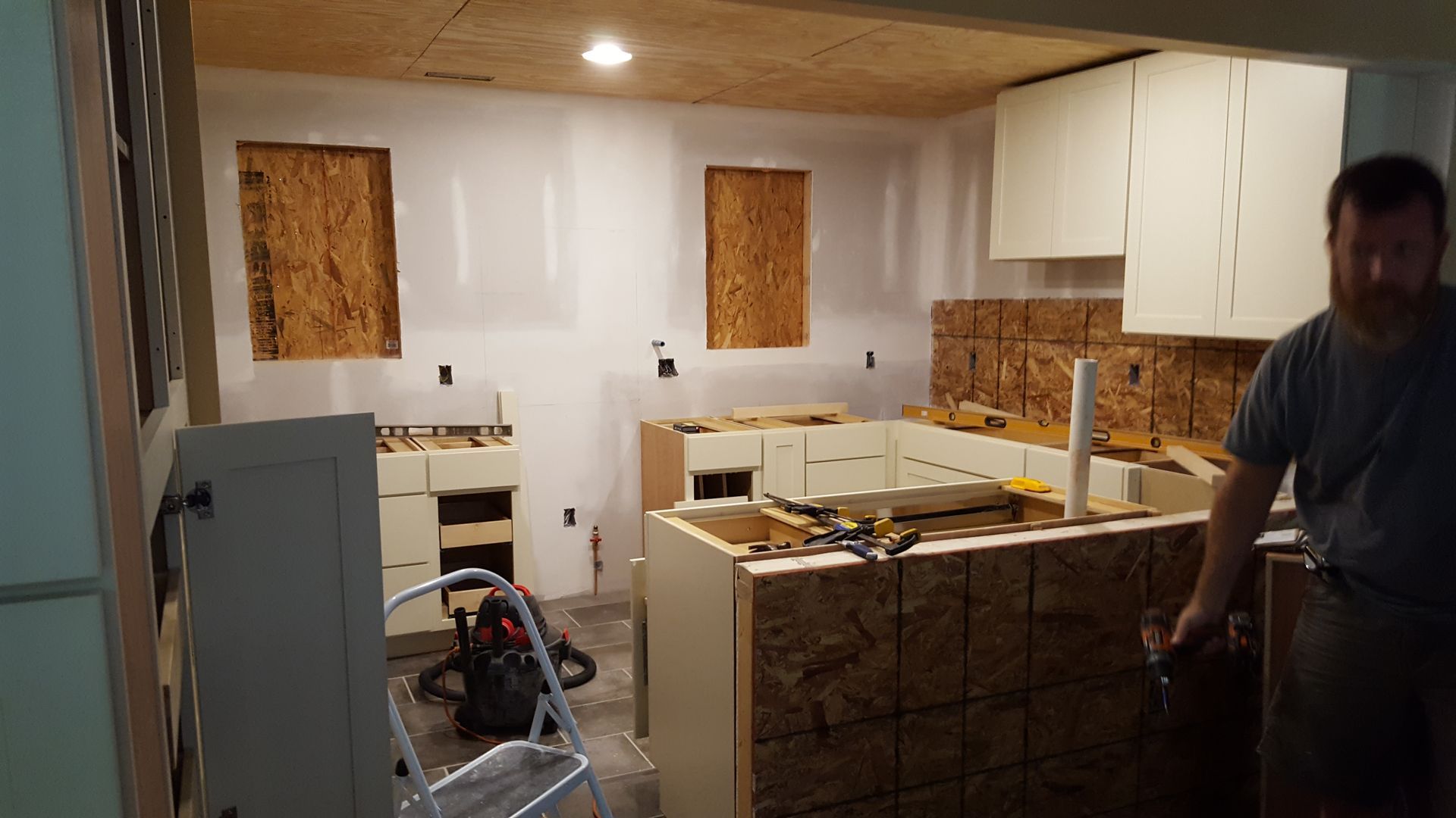 A man stands in a kitchen under renovation, with white cabinets, exposed wooden walls, and a ladder in the foreground.
