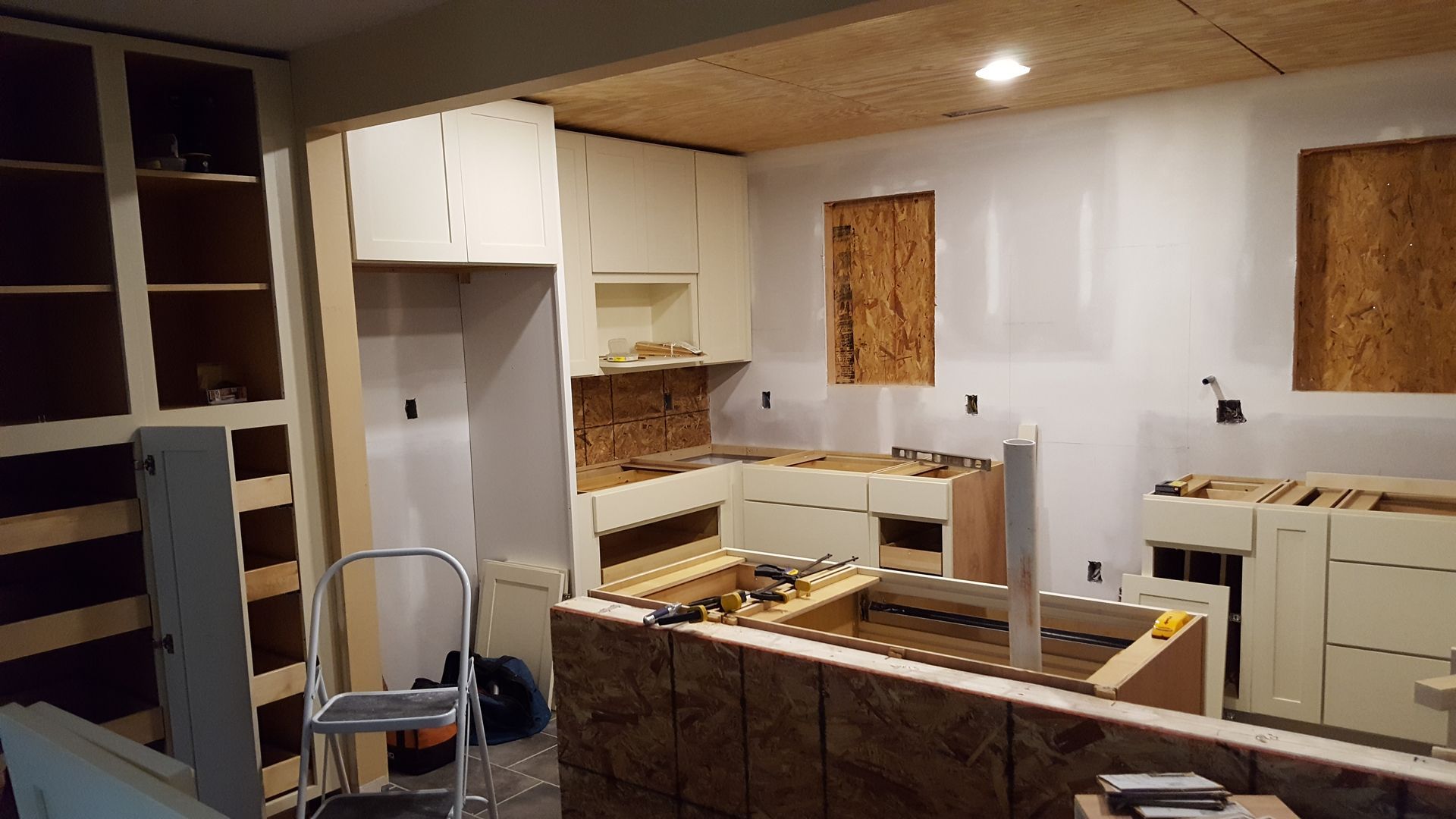 A kitchen undergoing renovation with unfinished cabinets, plywood walls and ceiling panels, and a step ladder.