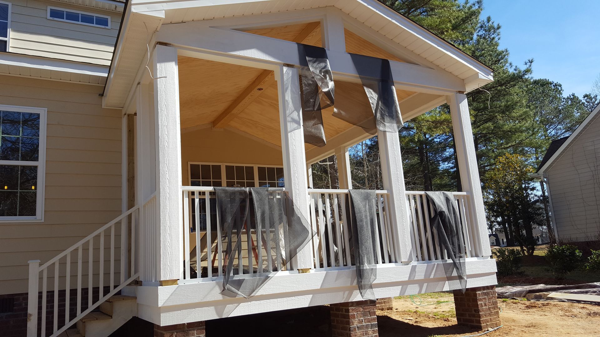 A tan house with a partially finished porch featuring white railings, pillars, and hanging construction mesh material.