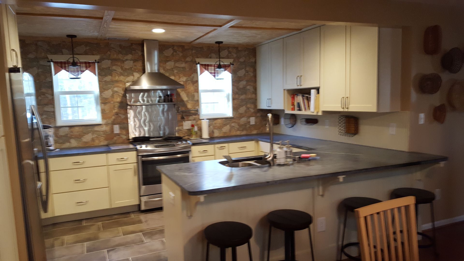 A rustic kitchen with cream cabinets, stone wall backsplash, stainless steel appliances, and a breakfast bar with stools.