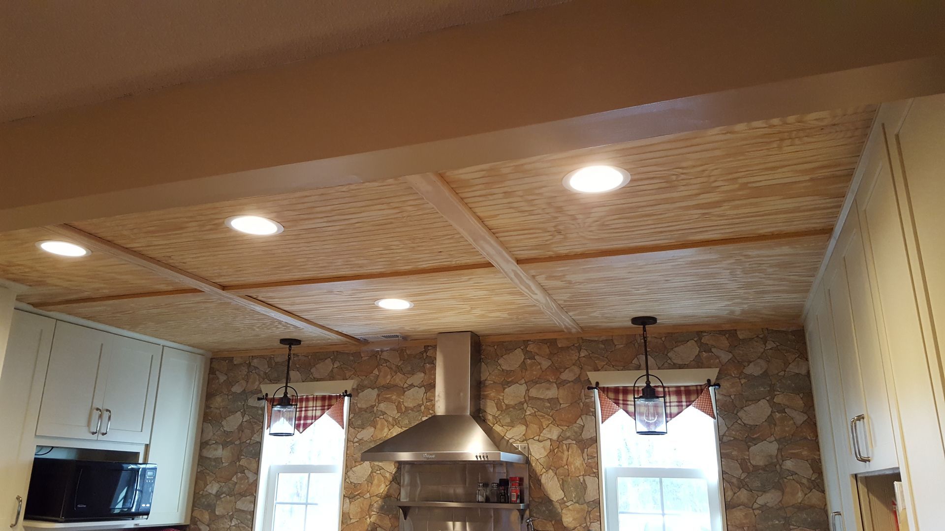 A kitchen ceiling with a light wood plank finish and recessed lighting, situated above a stone wall with a metal vent hood.