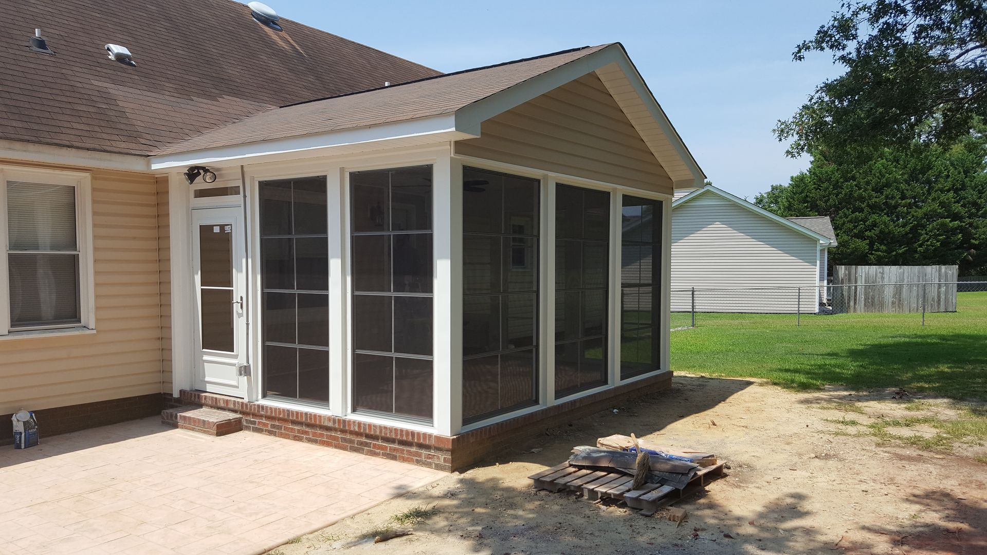 A newly built beige sunroom with screened windows and a brick base attached to the back of a house.