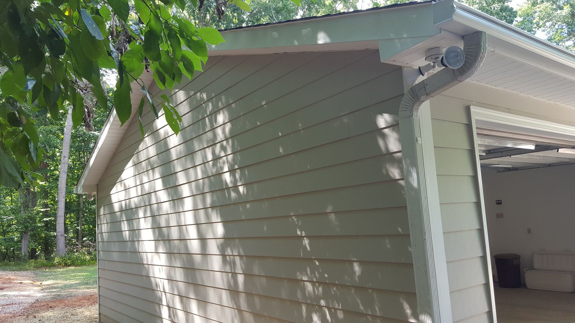 A side view of a light green garage with horizontal siding, a white gutter, and a partially open door near a tree line.