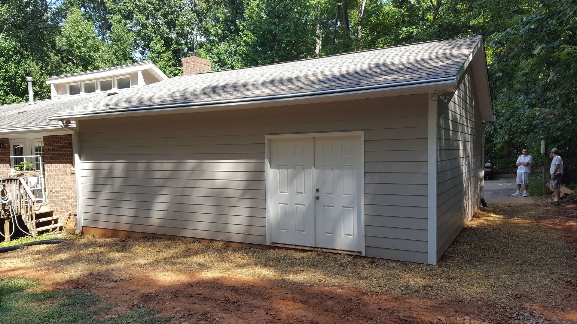 A beige, single-story garage addition with a gabled roof and double white doors, set against a wooded backdrop.