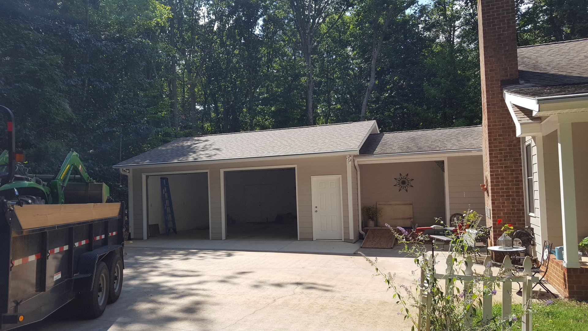 A tan-colored garage with two open bays and a side door, situated next to a house with a brick chimney and a white fence.