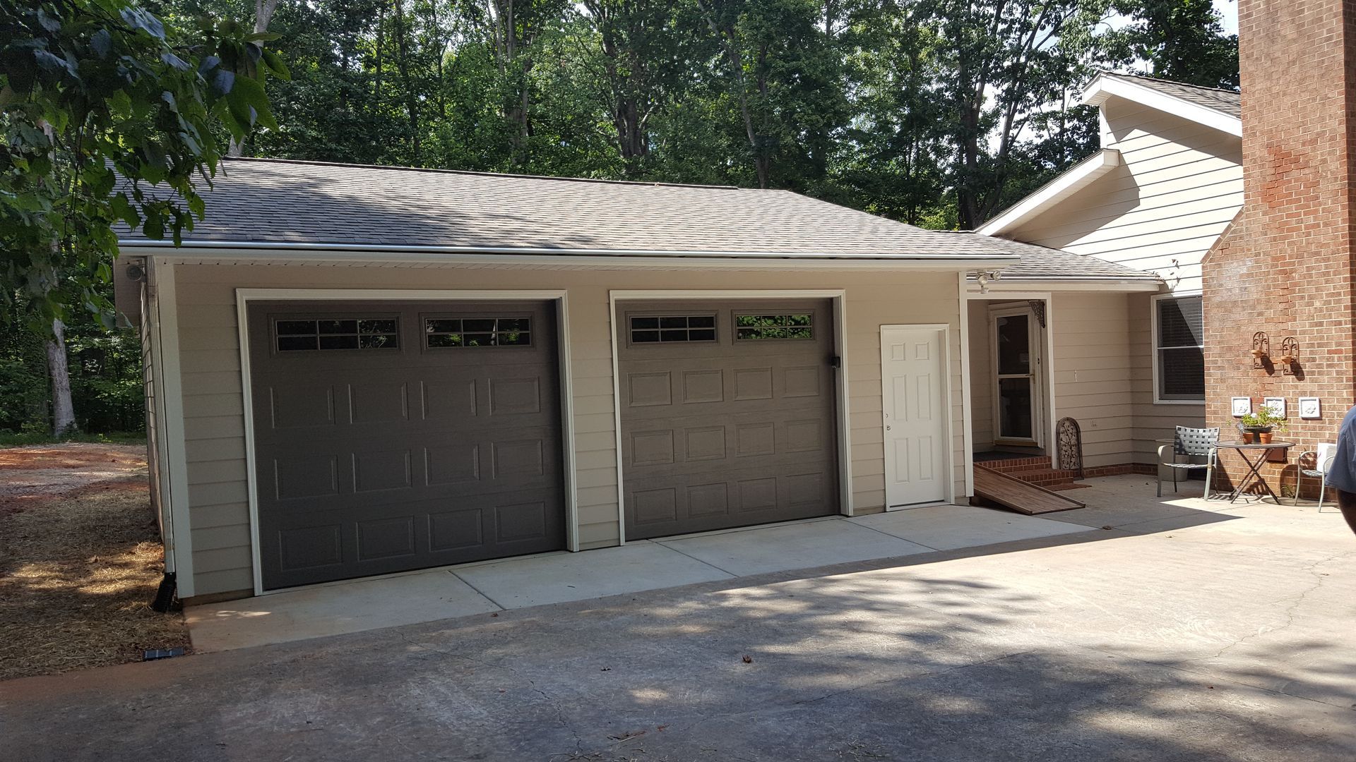 A tan two-car garage with brown doors, a white side door, and a shingled roof attached to a brick house.