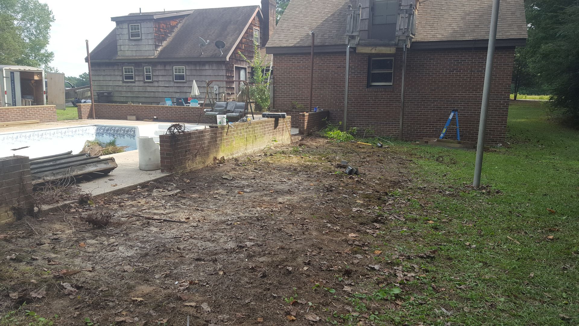A backyard landscape under construction, featuring a partially built pool, stone retaining walls, and two brick houses.