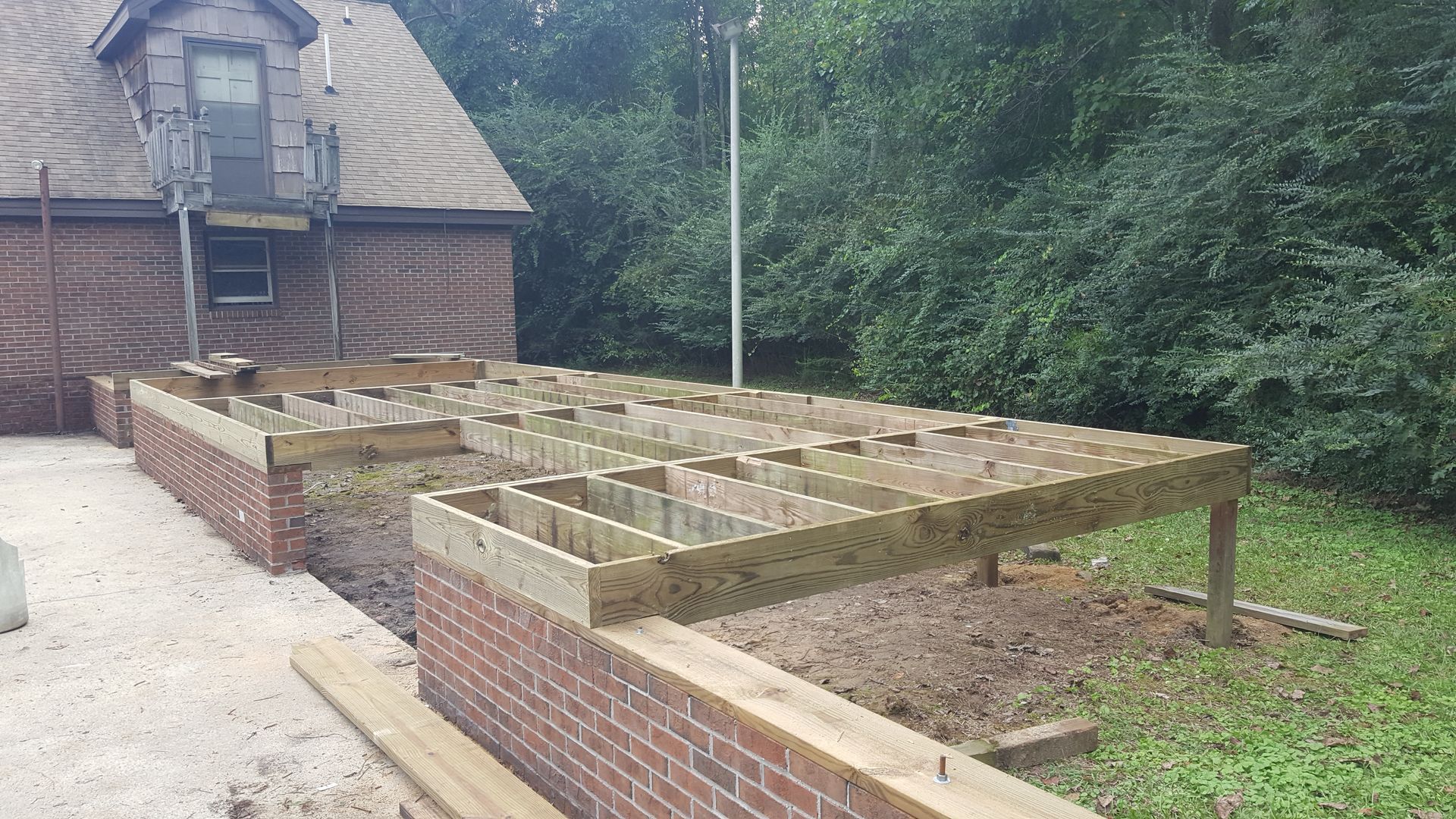 A wooden deck frame under construction atop a low brick wall next to a brick house in a backyard.