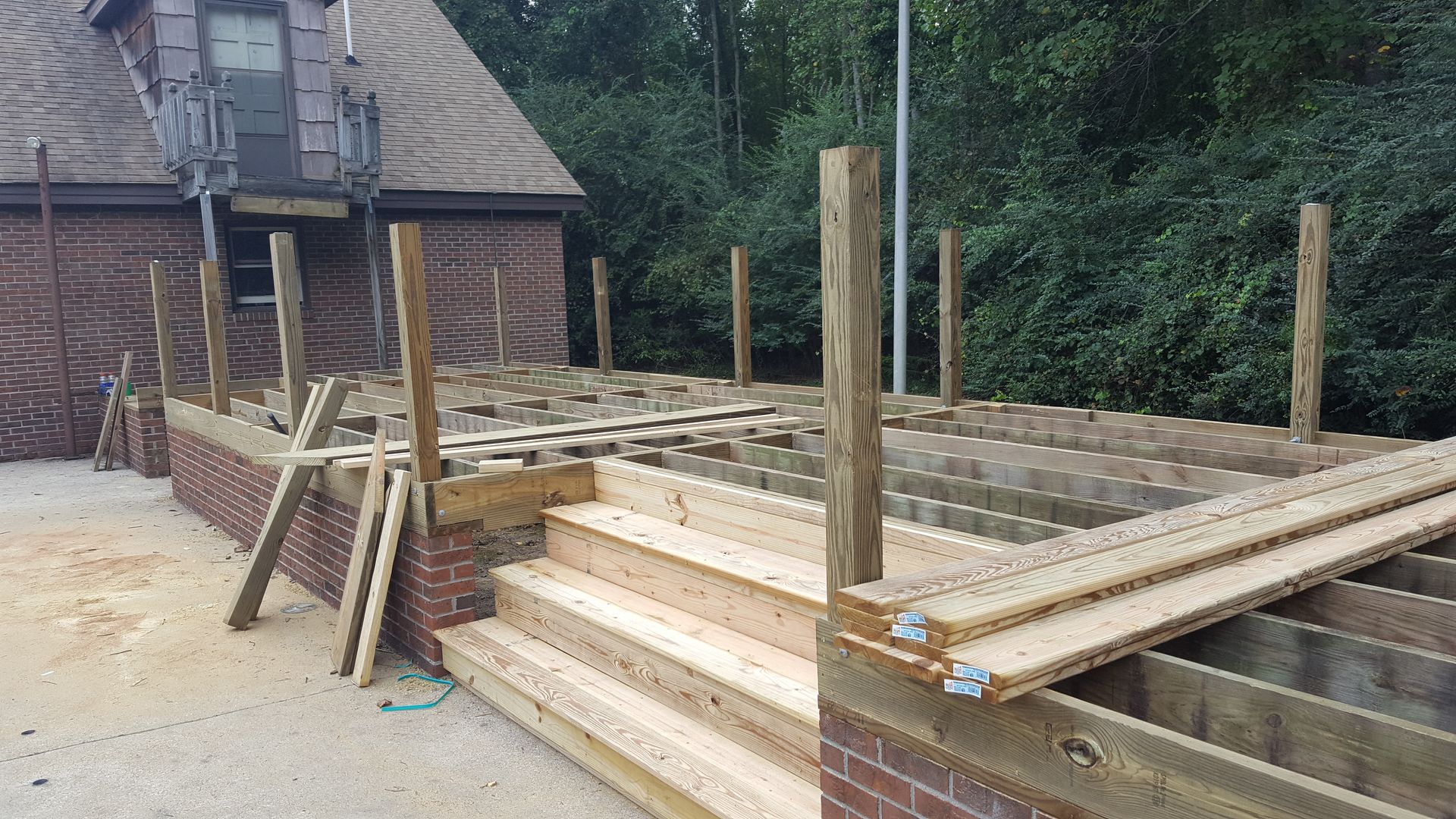 A wooden deck frame under construction featuring stairs leading up from a concrete patio beside a brick house.