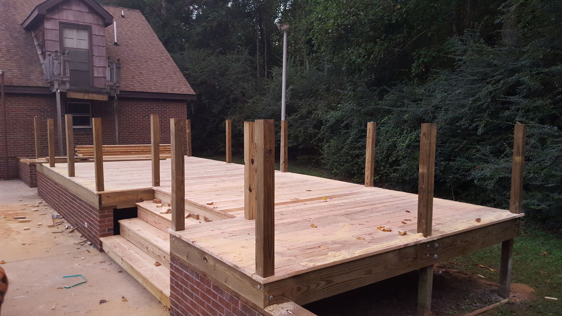 A wooden deck under construction with exposed posts and brick siding, set against a house and lush greenery.