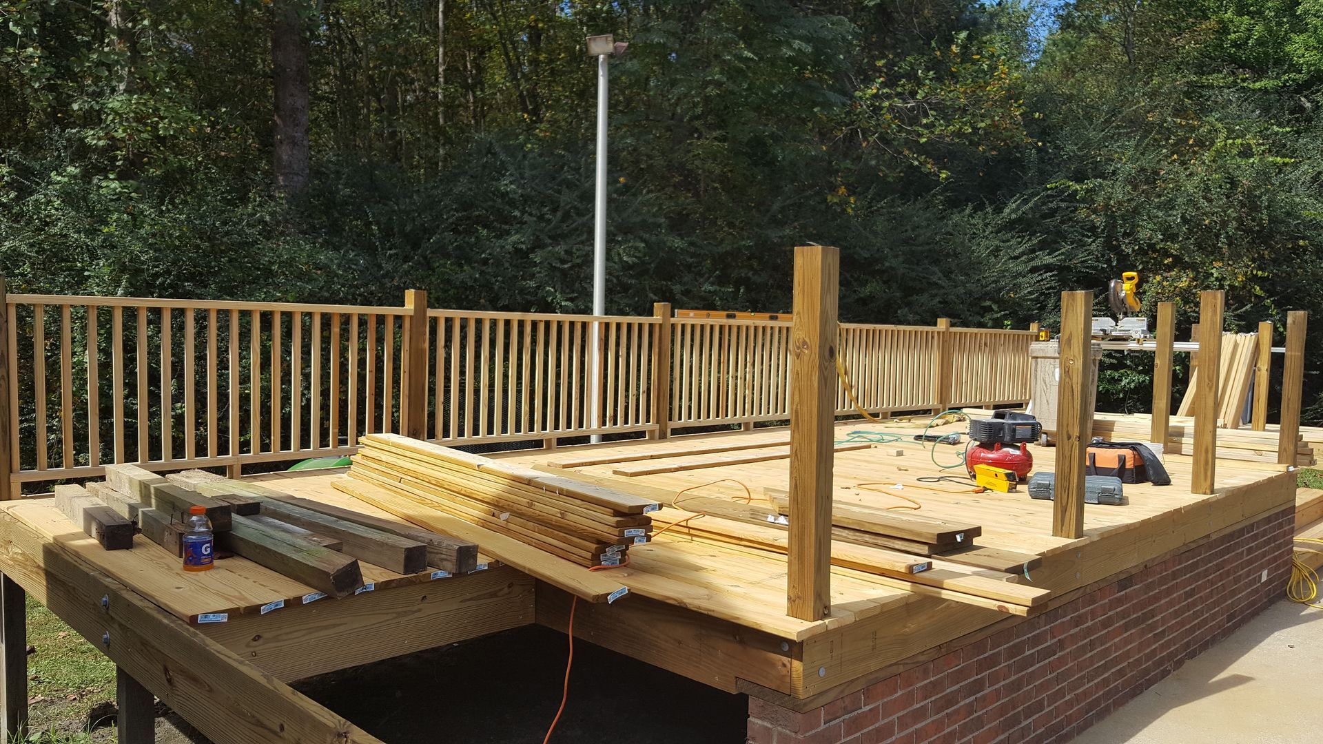 A wooden deck under construction with railings and tools, situated next to a brick building and lush green trees.