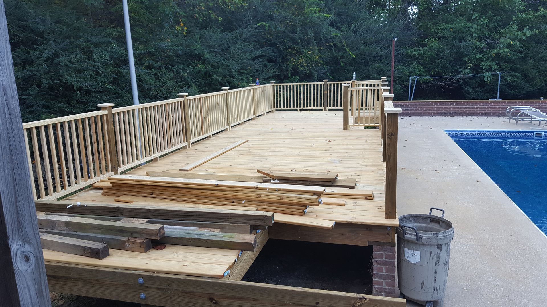 A wooden deck under construction next to a swimming pool, with piles of lumber and a trash can on the platform.