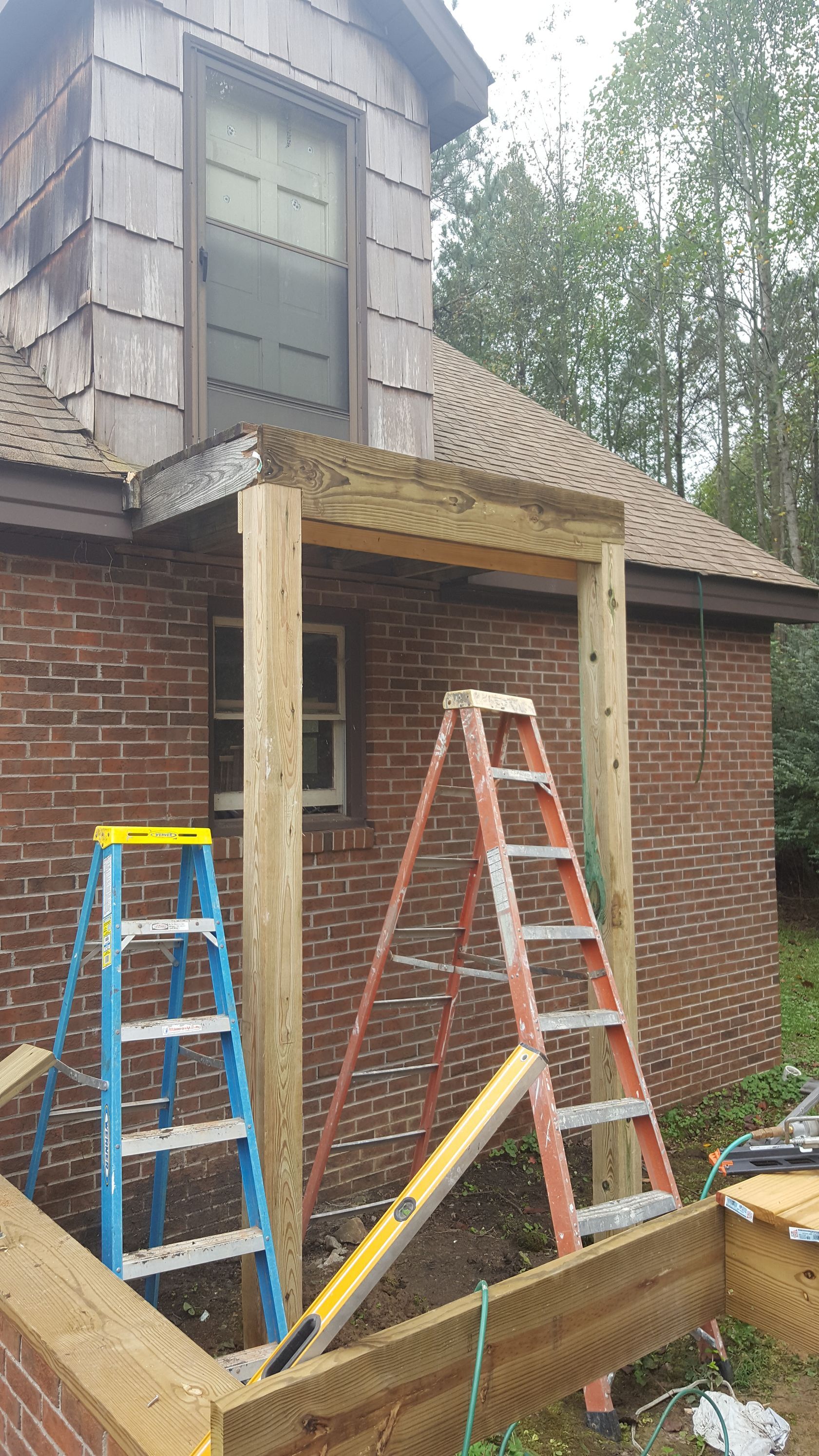 A construction site showing two wooden support posts being installed for a porch roof extension on a brick house.