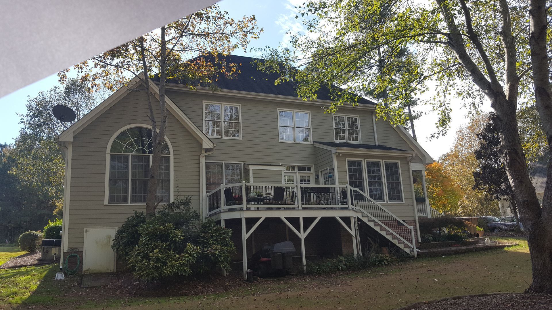 A two-story tan house with a deck, large windows, a dark roof, and surrounding trees on a sunny day.
