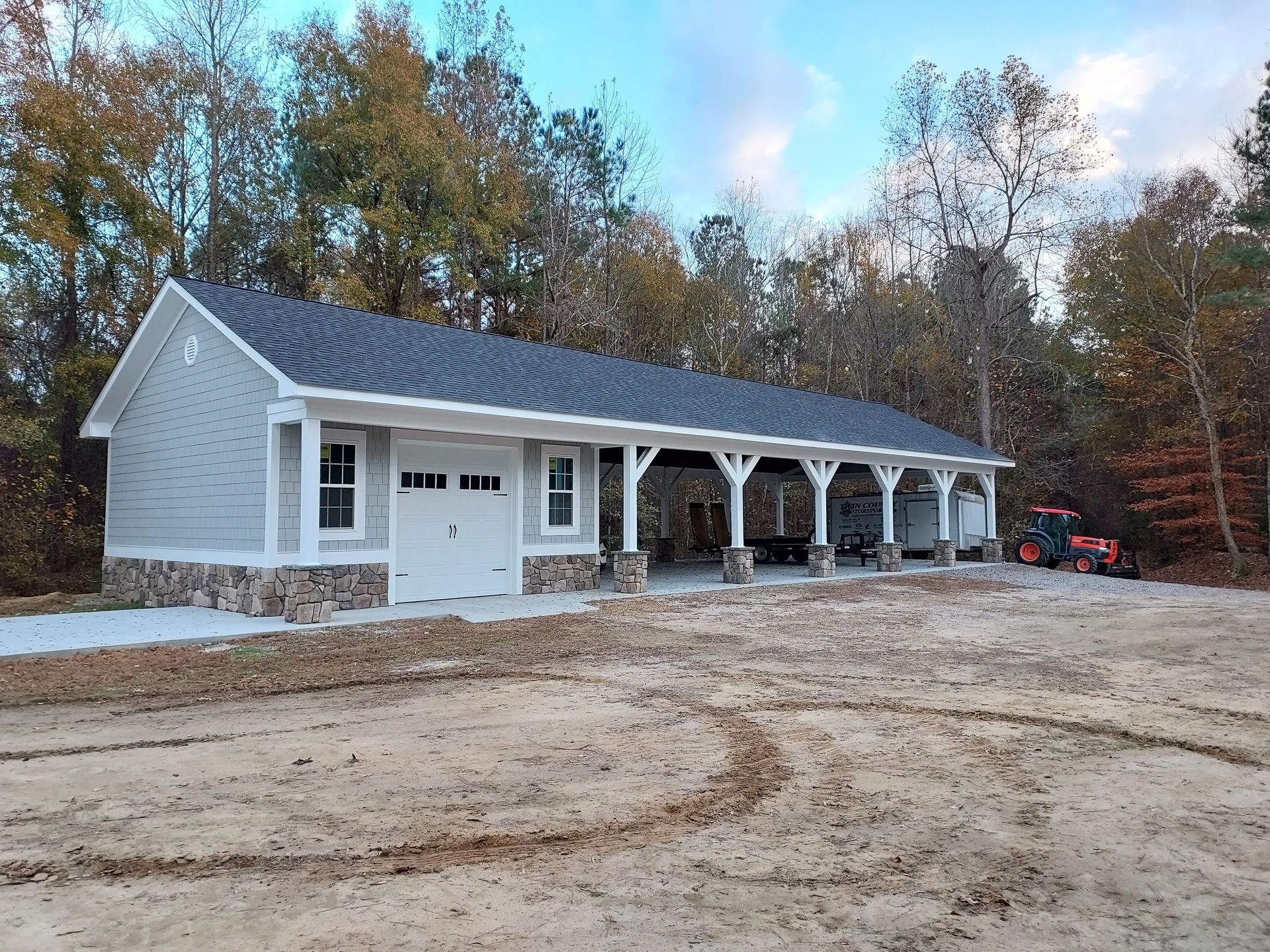A newly constructed light grey garage with a stone base and an attached open carport, set on a dirt lot near a forest.