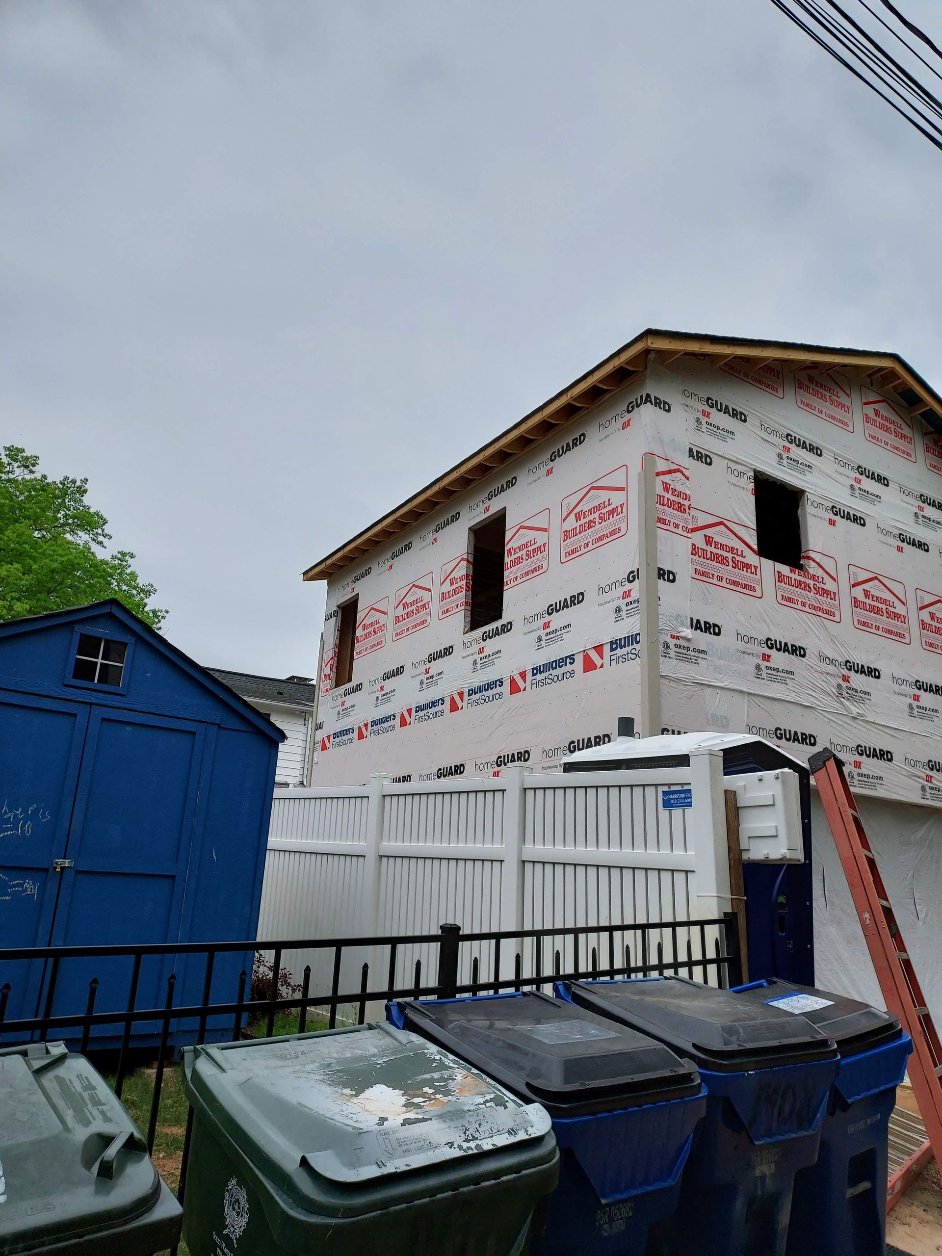 A two-story building under construction with white protective wrap, next to a blue shed, a white fence, and trash bins.