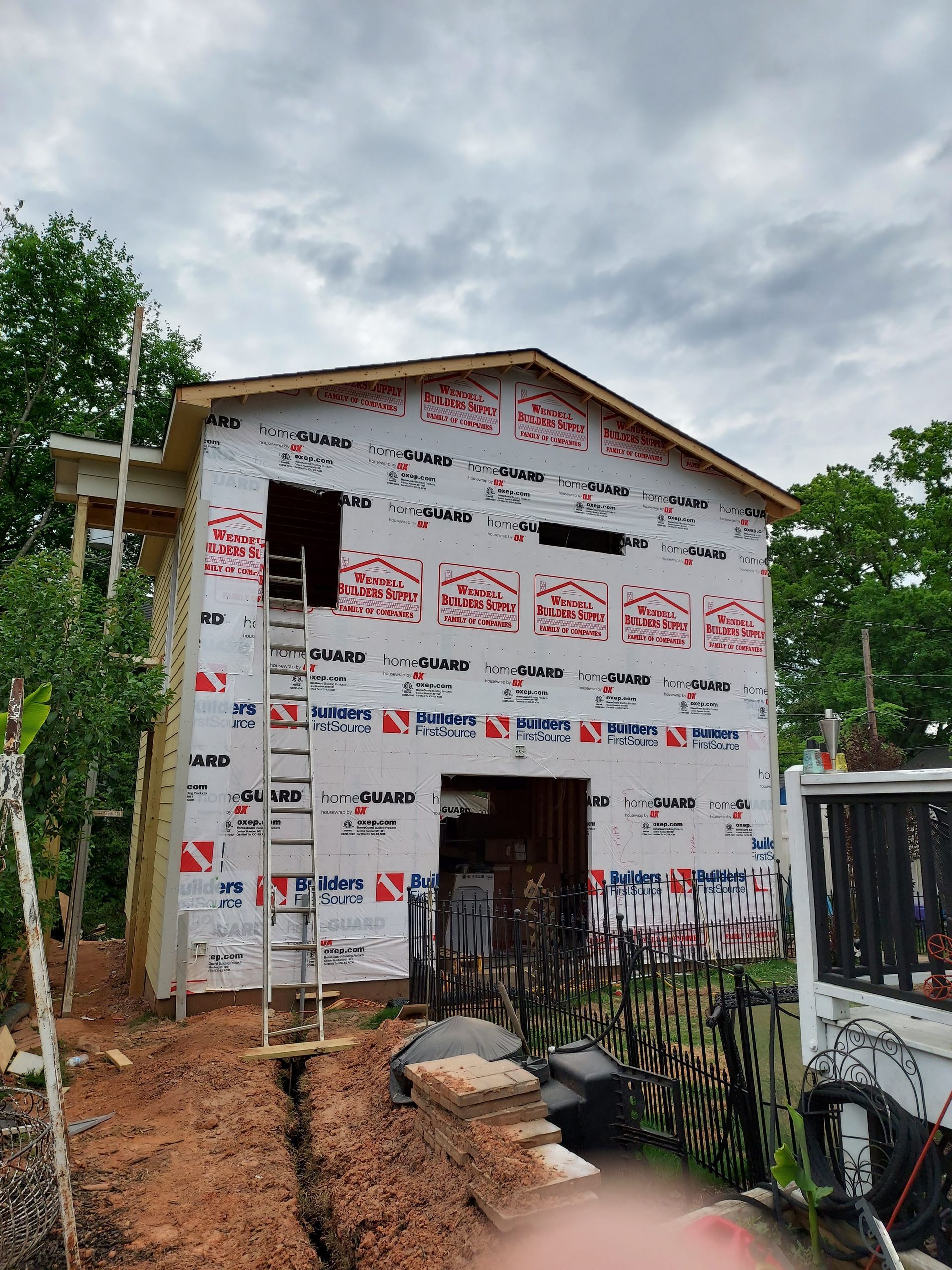 An unfinished two-story house under construction, wrapped in white protective sheeting with red logos, under a cloudy sky.