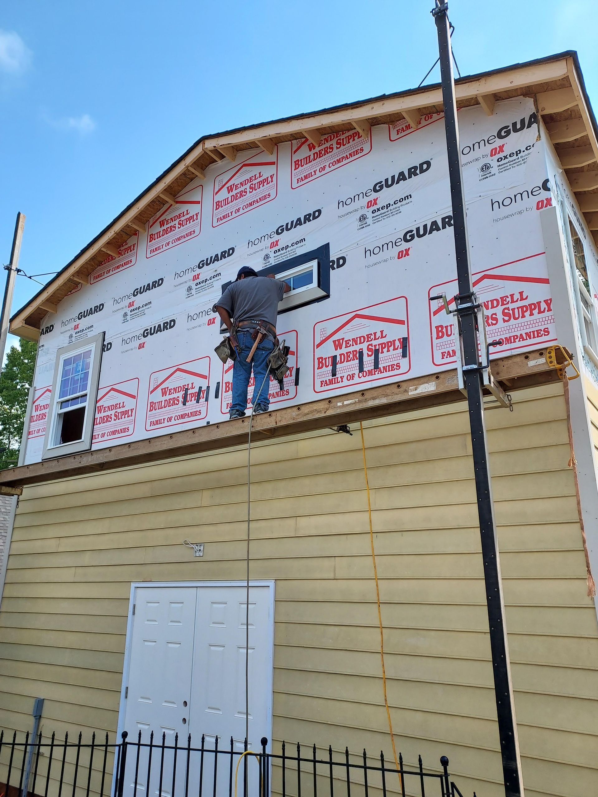 A worker installs a window on the upper story of a building exterior covered in red and white moisture barrier sheeting.
