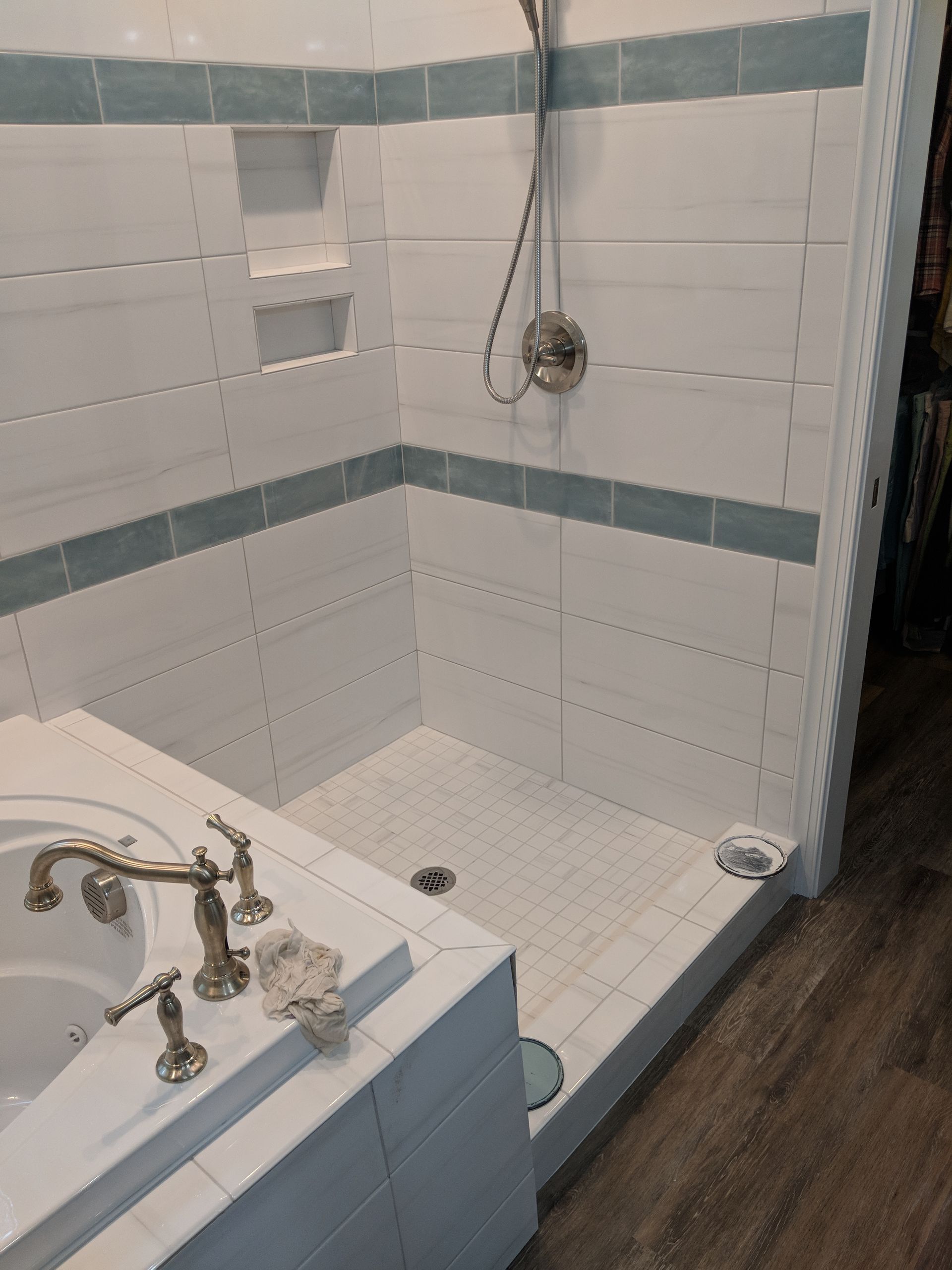 A tiled shower stall with a handheld shower head and two recessed shelves next to a white bathtub with gold fixtures.