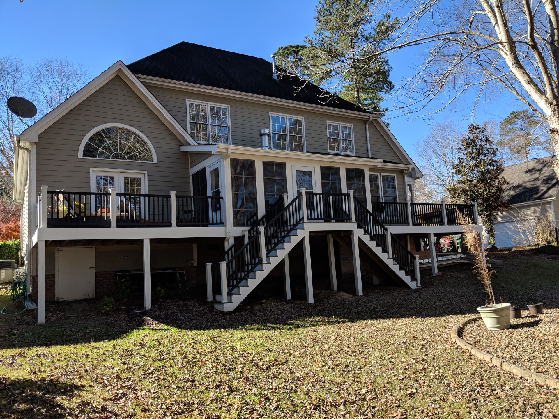 A two-story tan house with a dark roof, a screened-in porch, and a wrap-around deck with stairs leading to a grassy yard.