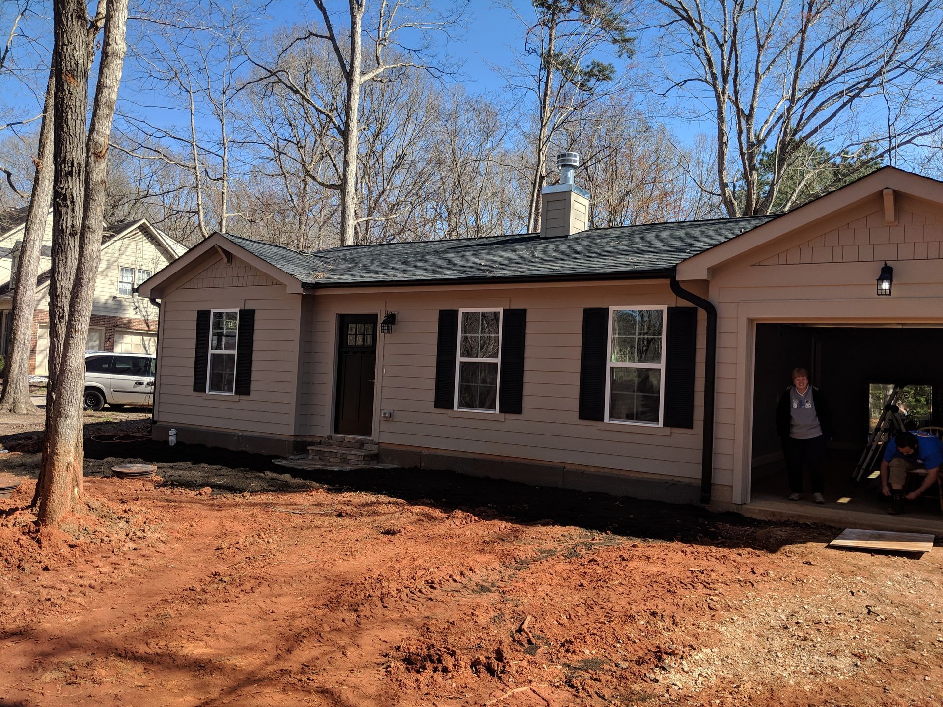 A single-story tan house with black shutters and a garage under a clear blue sky, set against a backdrop of bare trees.