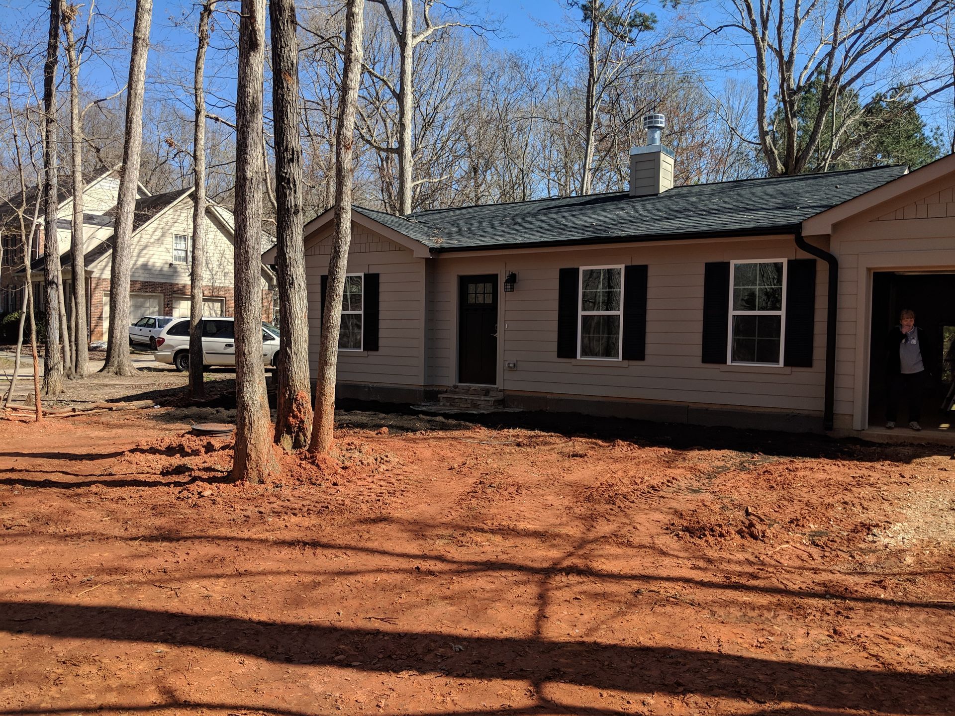 Single-story beige house with a dark roof and black shutters, situated behind a patch of bare, red clay ground.