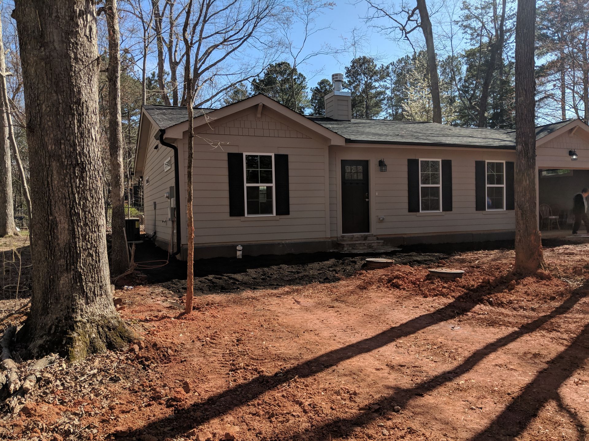 A one-story, light-colored house with black shutters and a front door sits in a wooded lot with reddish soil.