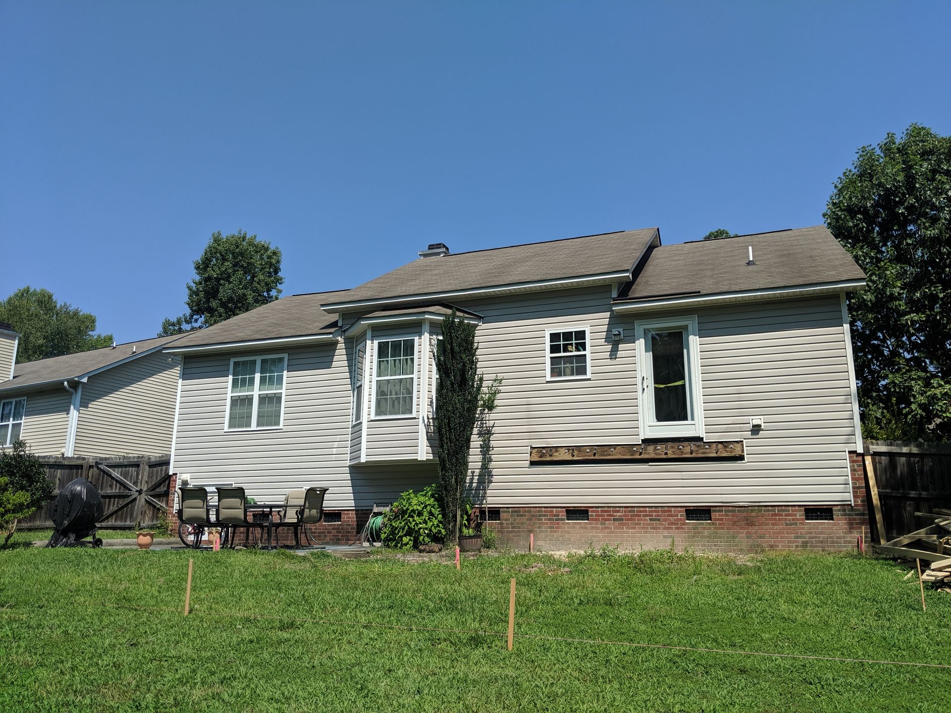 A single-story house with gray horizontal siding, a bay window, and a brick foundation viewed from a grassy backyard.