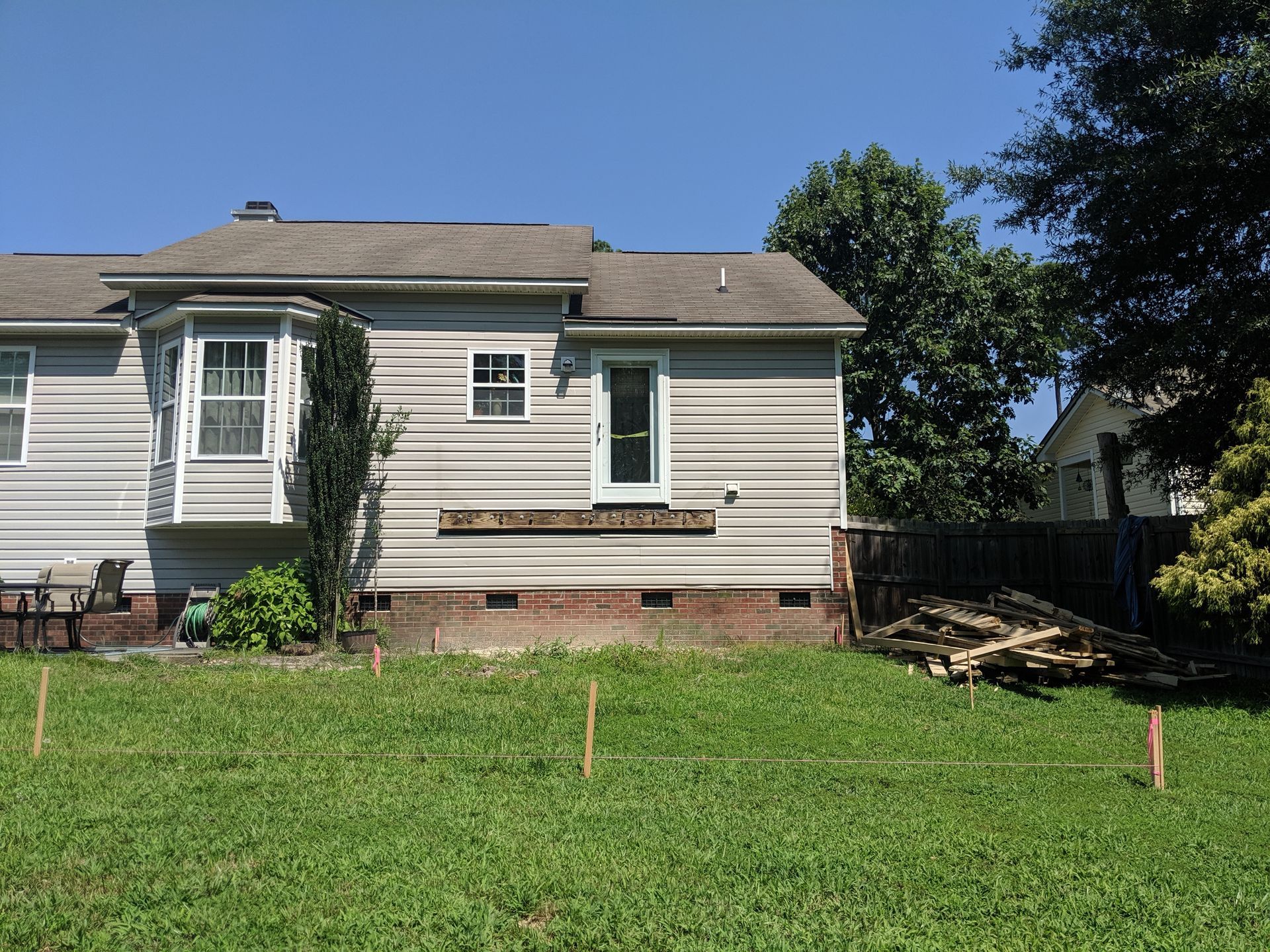 A light gray house with a brick foundation sits behind a grassy yard featuring wooden stakes in the ground.