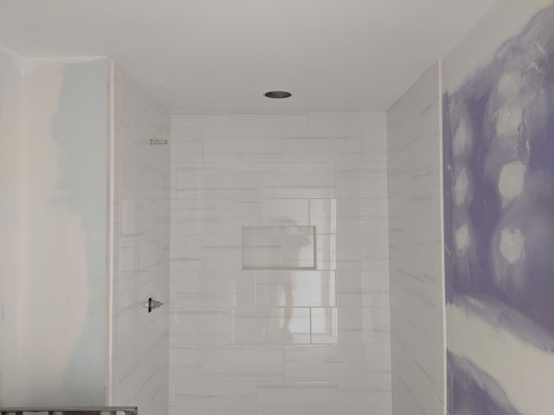 A shower alcove with white tiled walls, a built-in recessed shelf, and unfinished purple and white drywall on the sides.