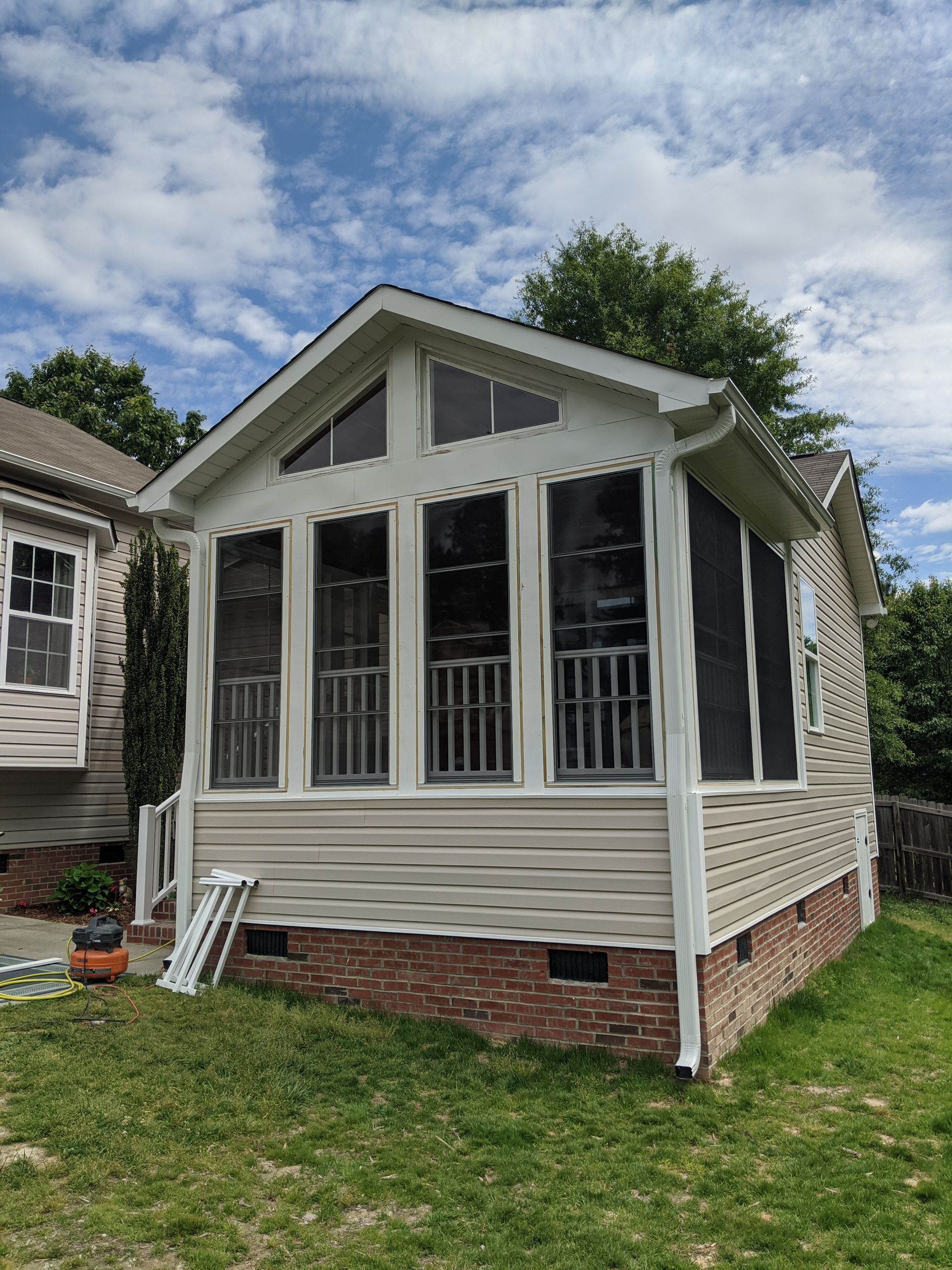 A light-colored sunroom addition with large windows and a brick foundation sits on a grassy lot under a cloudy sky.