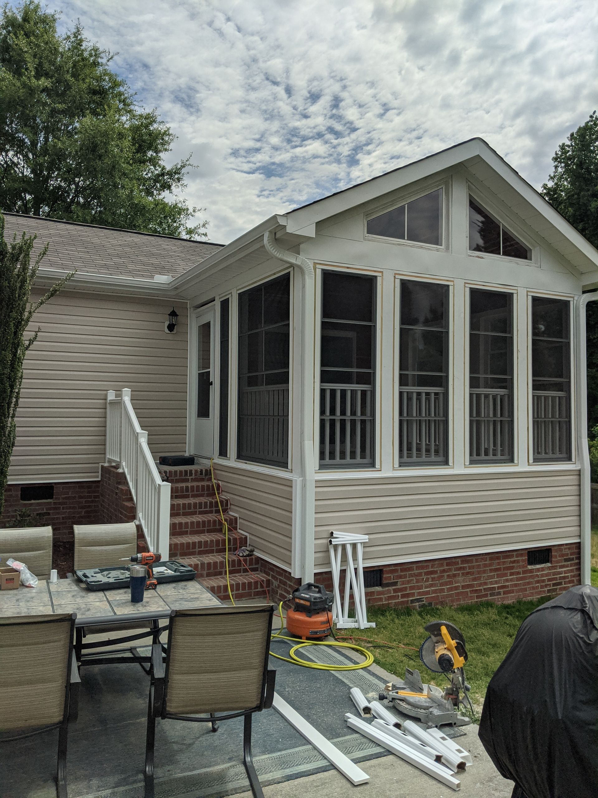A beige sunroom addition on a house with a brick foundation, stairs, and patio furniture in the foreground.
