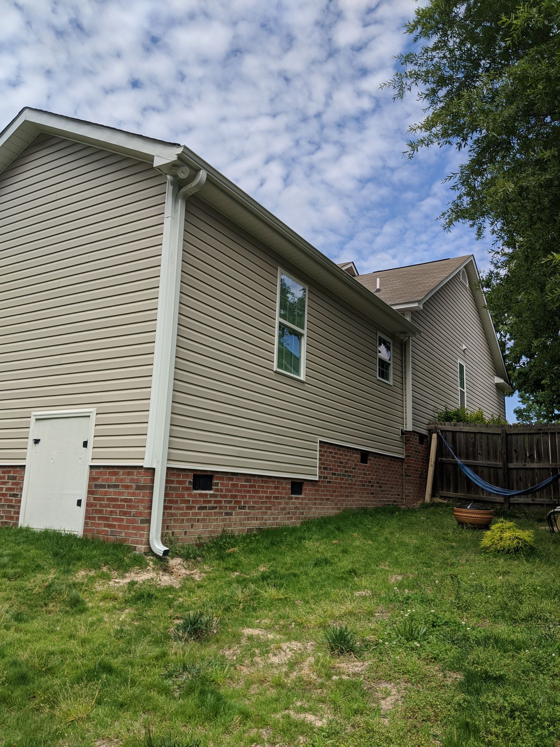 A beige-sided house with a brick foundation and a white door, set against a cloudy blue sky and a grassy yard.