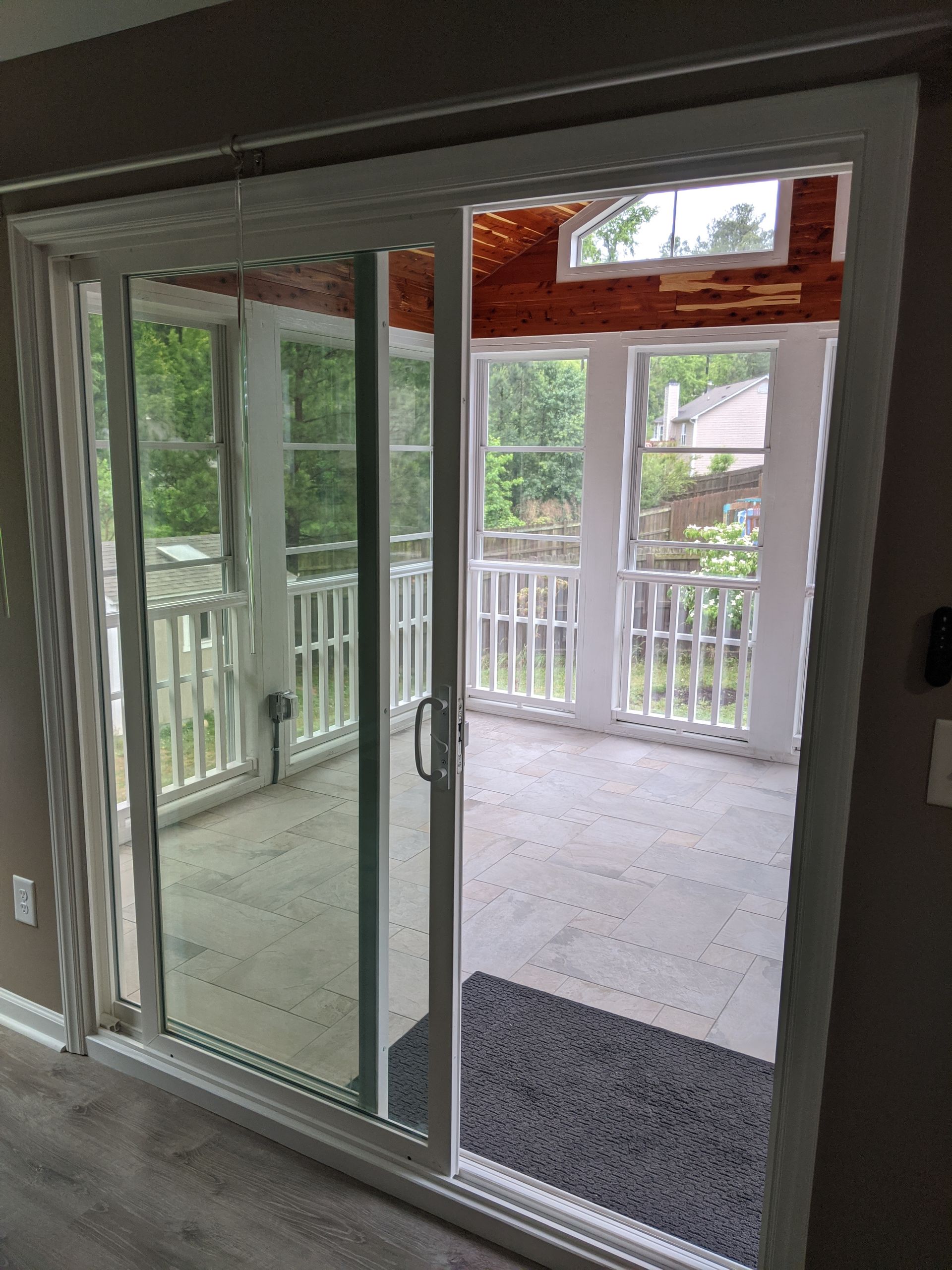 A white sliding glass door opens to a bright, tiled sunroom with white railings and trees visible through the screens.