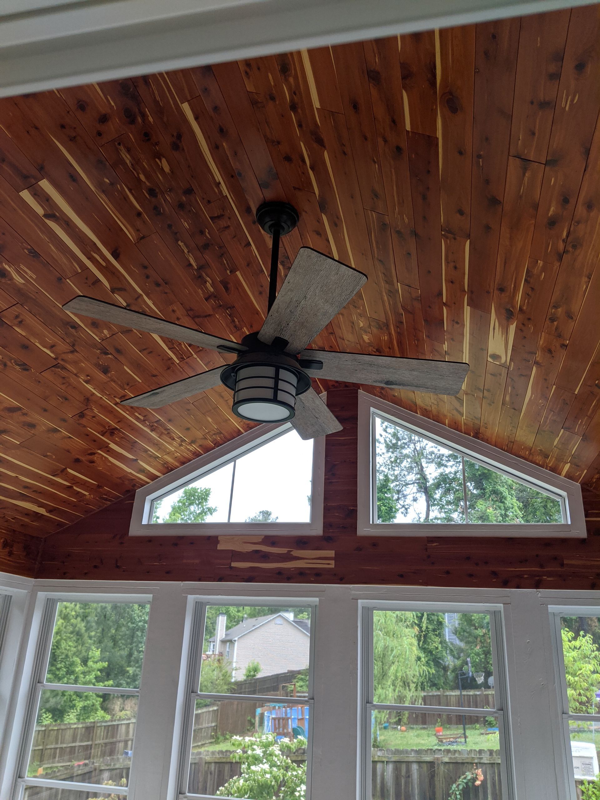 A ceiling fan with rustic gray blades hanging from a wood-plank ceiling above windows looking out onto a backyard.