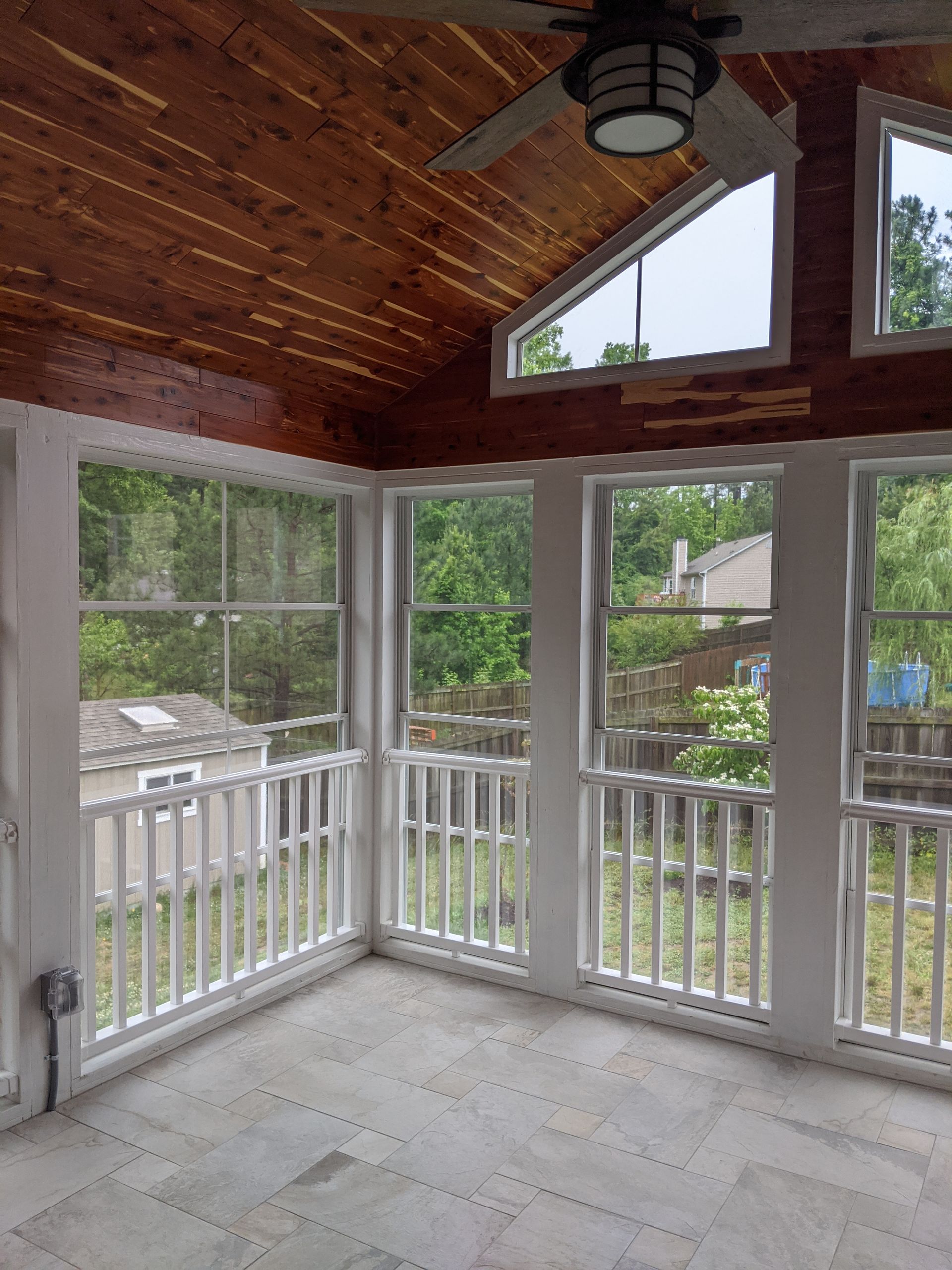 A sunroom with white railings, grey tile flooring, and a slanted wood-paneled ceiling featuring a mounted ceiling fan.