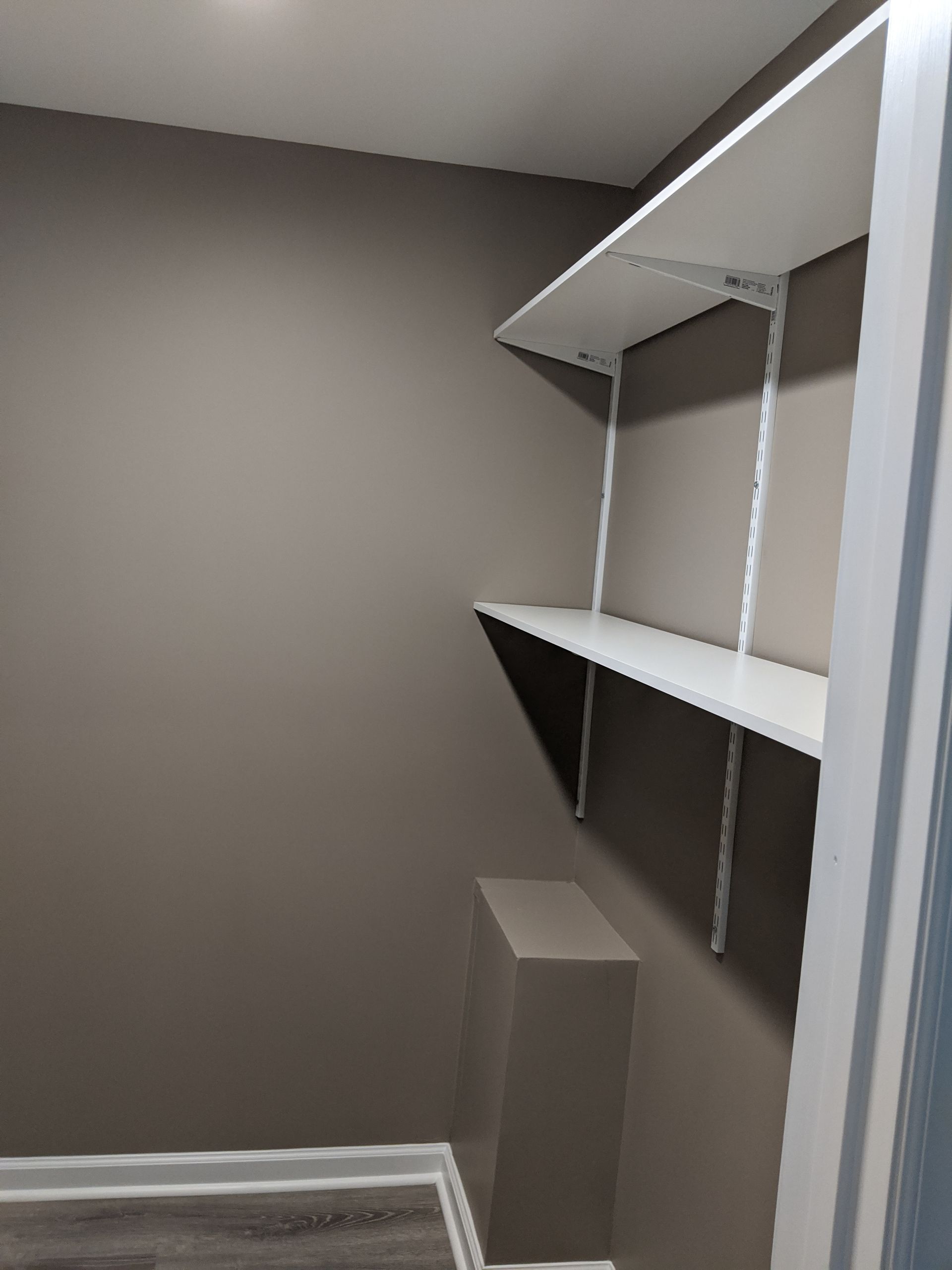Two white adjustable shelves mounted on a light brown wall above a small matching enclosure in a closet.